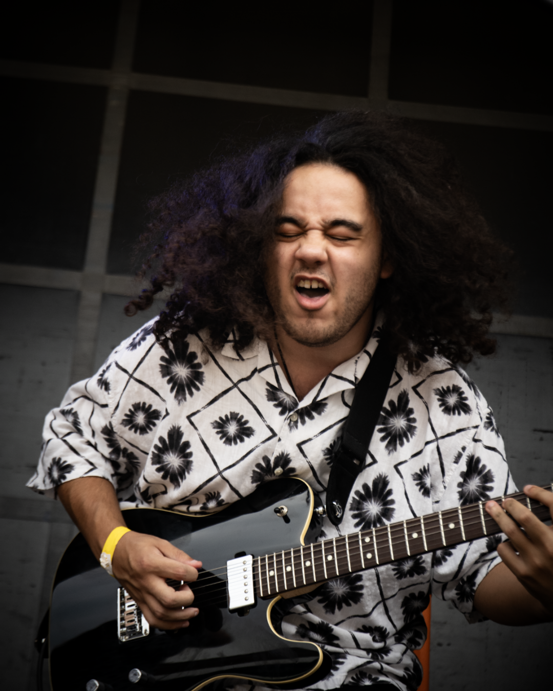A man with long curly hair playing an electric guitar, wearing a floral patterned button-up shirt, with a passionate expression.