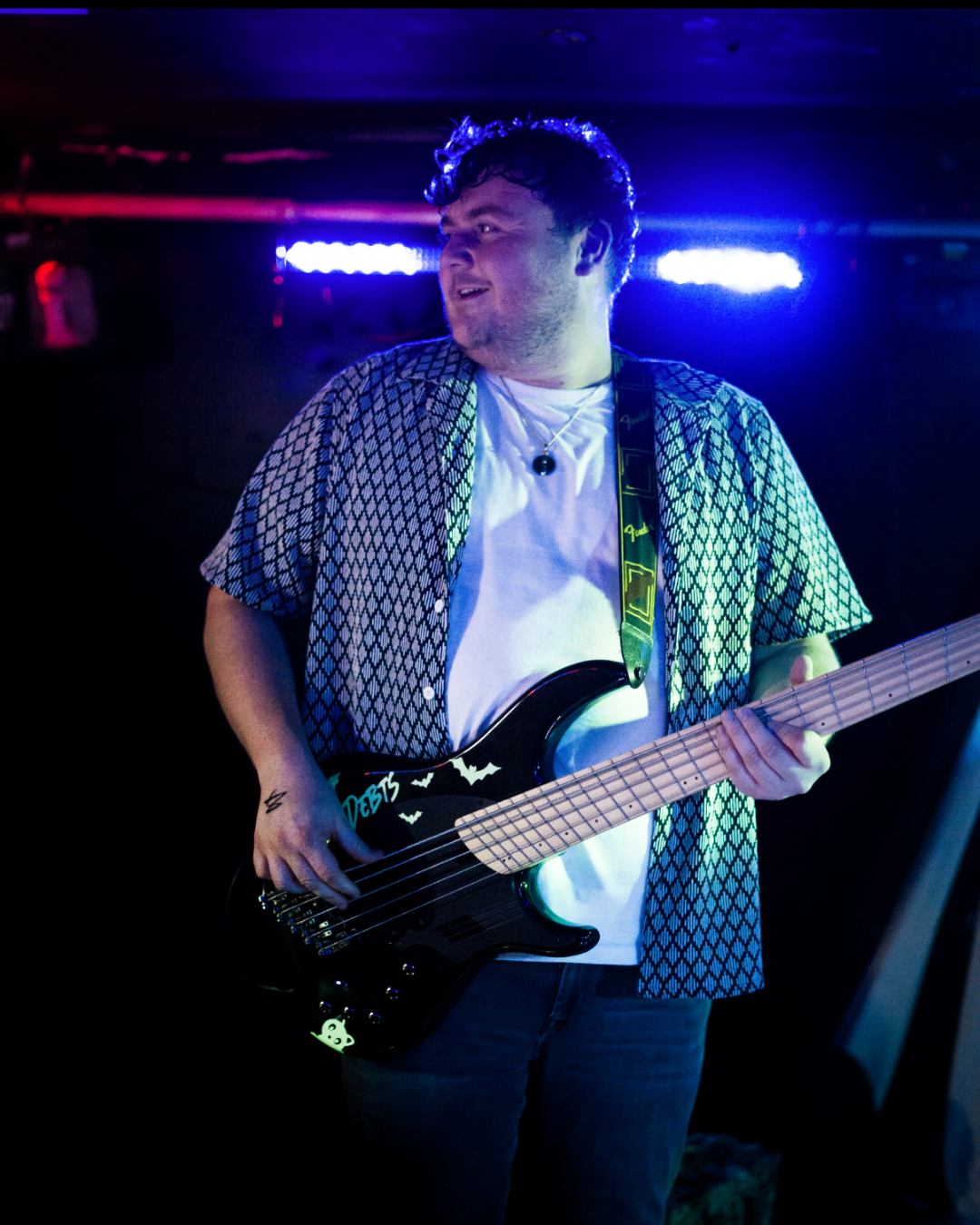 A man playing a guitar on stage, illuminated by purple and blue stage lights, wearing a patterned short-sleeve shirt over a white T-shirt, with a pendant necklace.