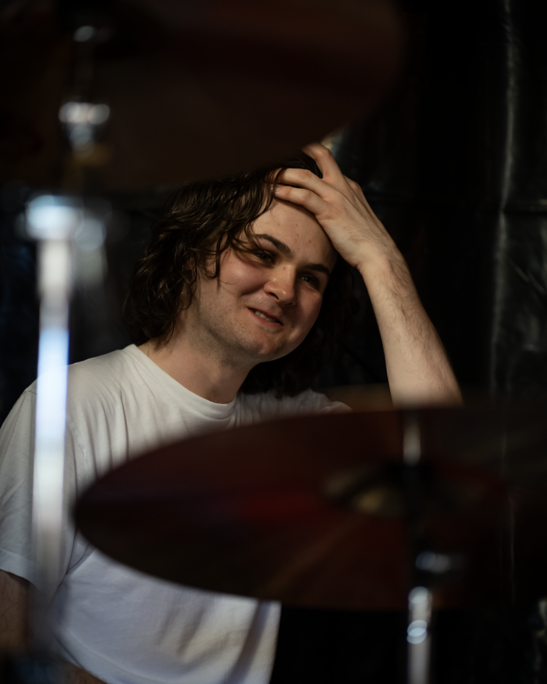 A young man with curly brown hair, wearing a white t-shirt, smiling, touching his forehead with his right hand, surrounded by musical instruments like cymbals, in a dimly lit setting.