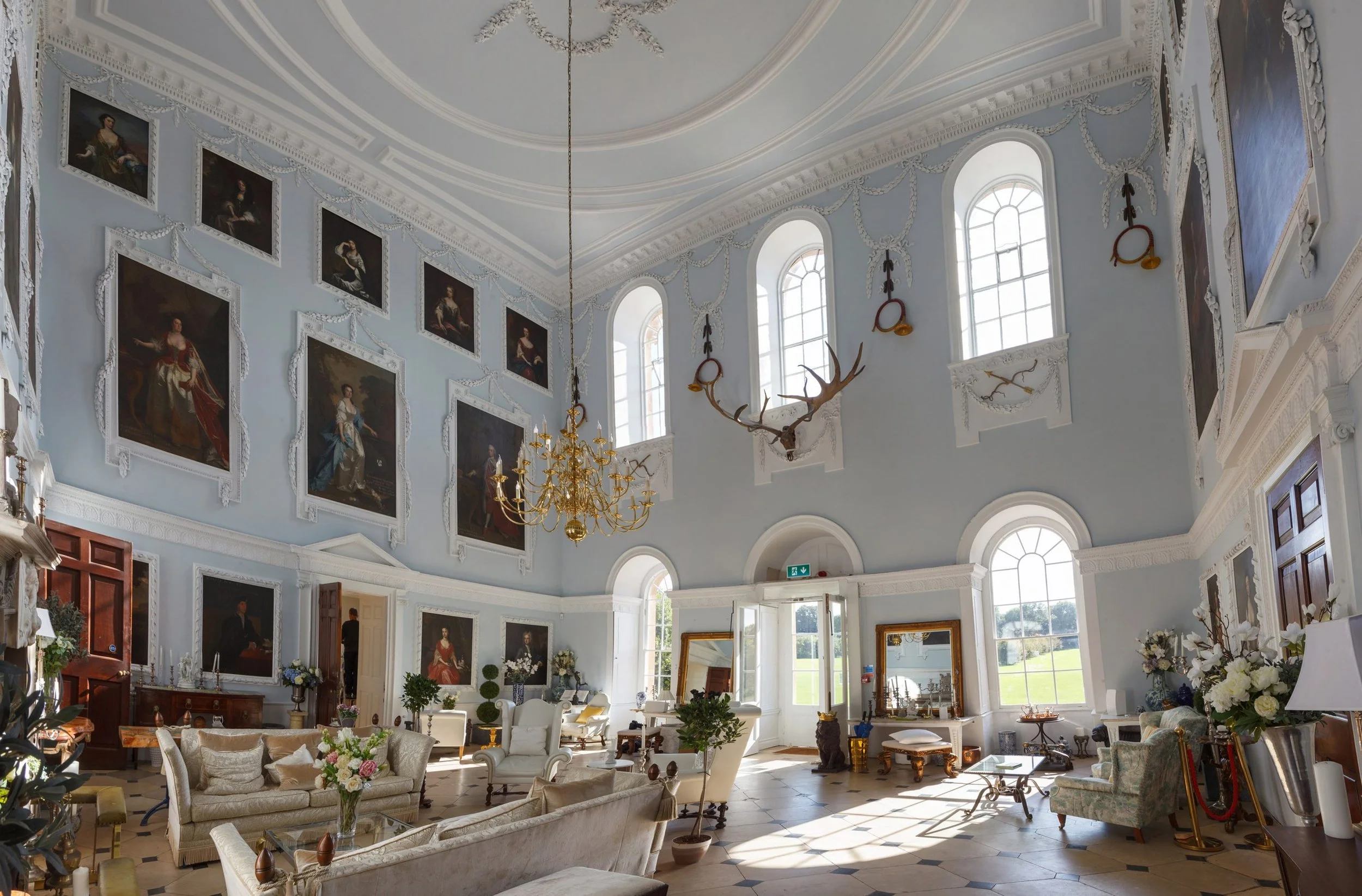 Interior of a spacious, elegant room with high vaulted ceiling, large arched windows, and ornate white decor. The room is furnished with sofas and armchairs, decorated with flowers and artwork, including a mounted deer head and numerous framed portraits on the walls. A chandelier hangs from the ceiling.