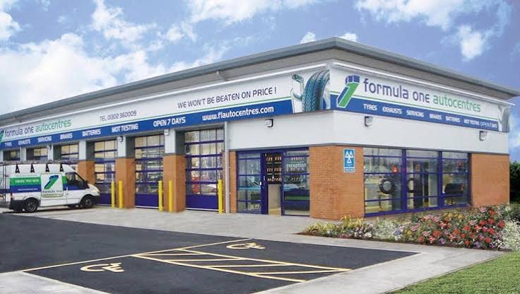 Exterior view of Formula One Autocentres with a white company van parked in front and a designated handicap parking space in the foreground. The building has large windows, a brick wall section, and signs indicating tire, exhaust, suspension, brakes, batteries, and vehicle testing services.