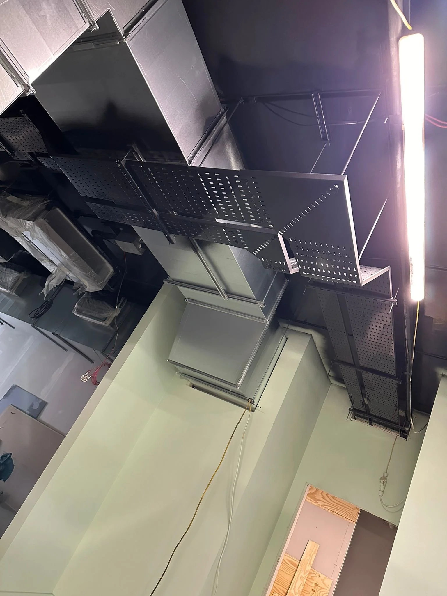 Construction site interior with black metal framing and a pale green wall, exposed electrical wiring, and a partially removed wooden floor, under construction lighting.