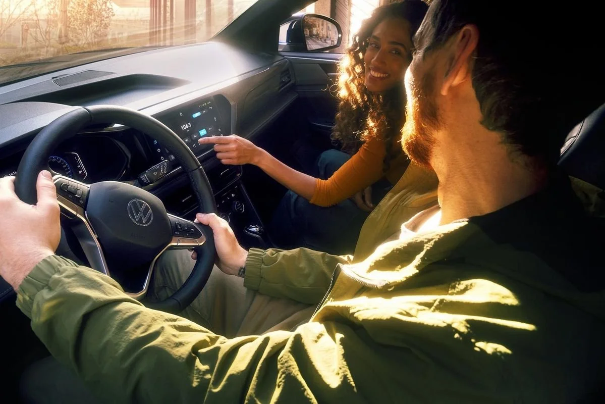 Un homme et une femme dans une voiture, le homme conduit et la femme pointe un écran tactile au tableau de bord, tous deux souriants, lors d'une journée ensoleillée.