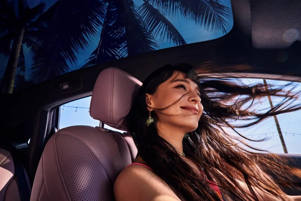 Une femme souriante avec les cheveux au vent à l'intérieur d'une voiture, contre un décor de palmiers et un ciel bleu au crépuscule.