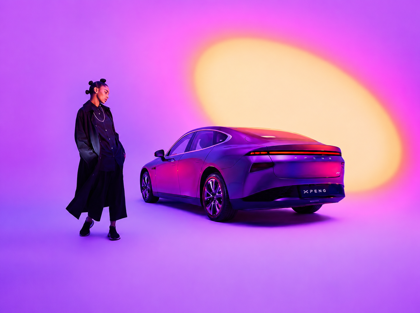 Une femme habillée en noir se tient à côté d'une voiture électrique violette dans un studio avec un fond coloré en dégradé de violet, rose et jaune.