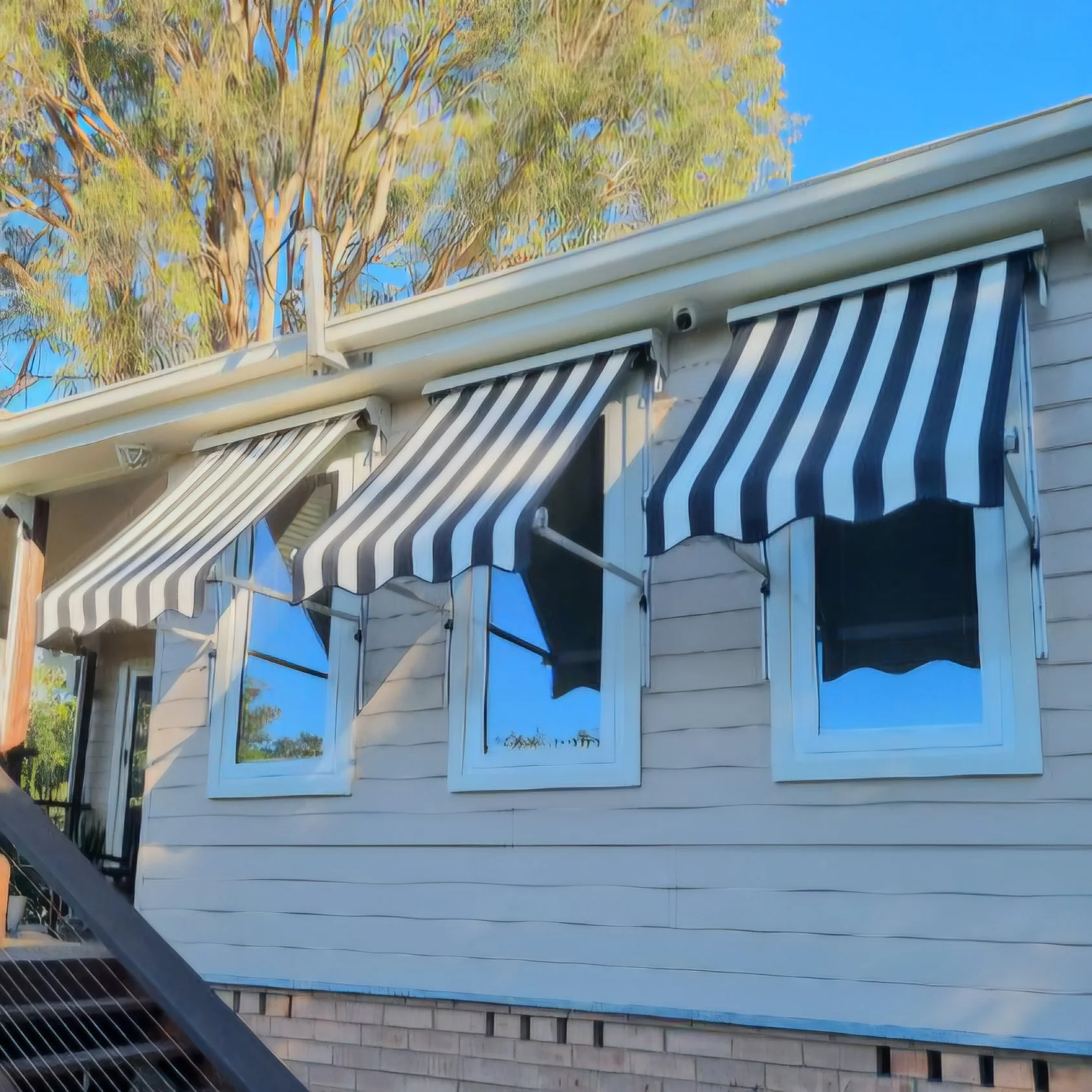 A house with multiple windows and striped awnings, with tall trees and a clear blue sky in the background.