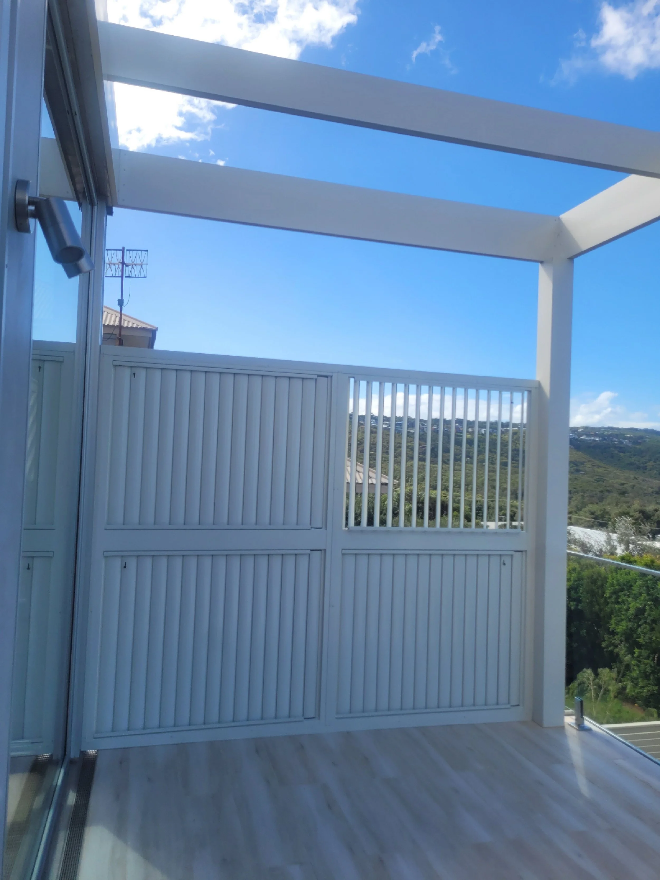 View from a balcony with white railings, a glass door, and a roof structure, overlooking a landscape of trees and hills under a blue sky with some clouds.