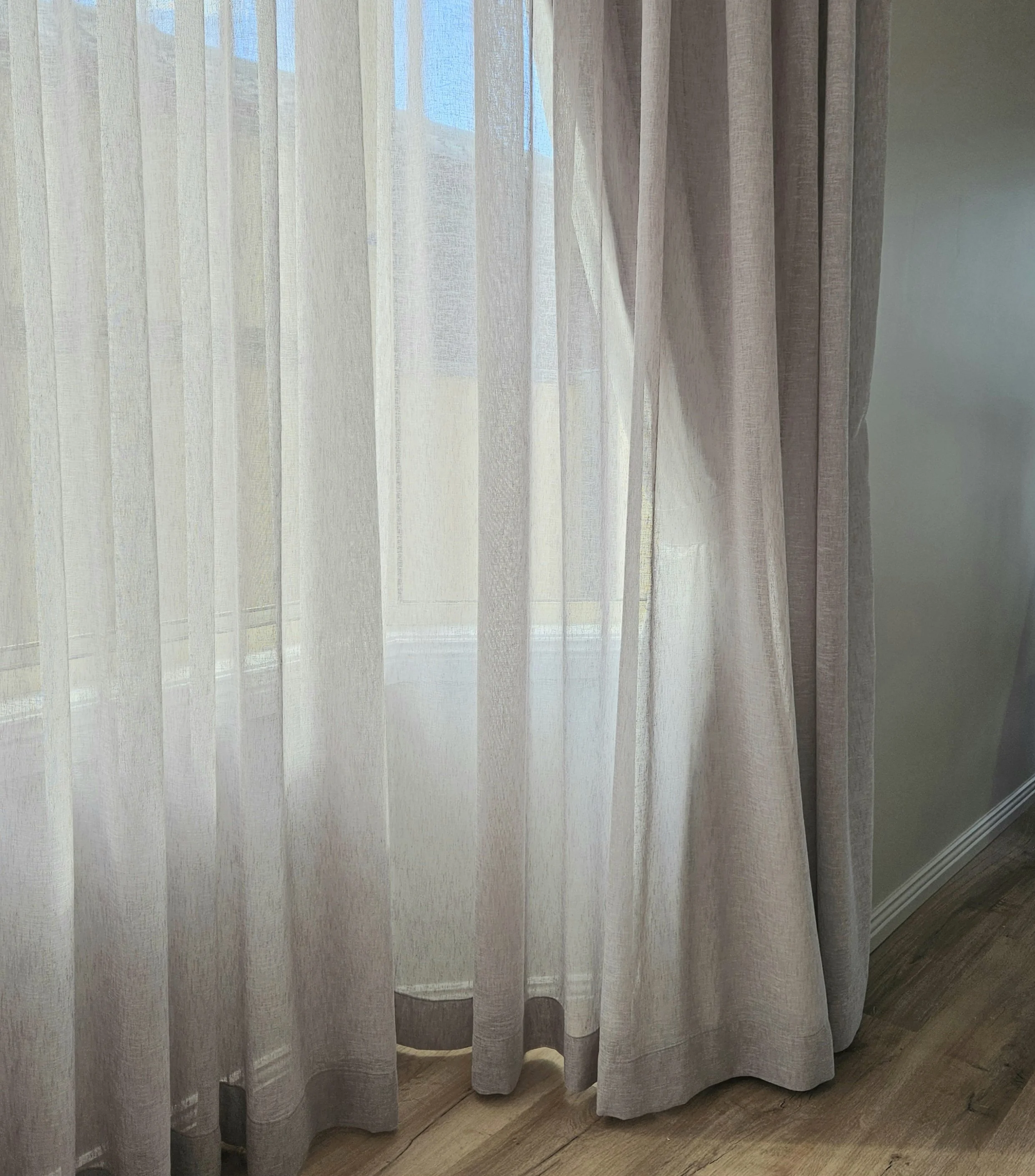White sheer curtains hanging in front of a window, with sunlight filtering through, covering a glass window and a view of the outside sky and part of a neighboring building.