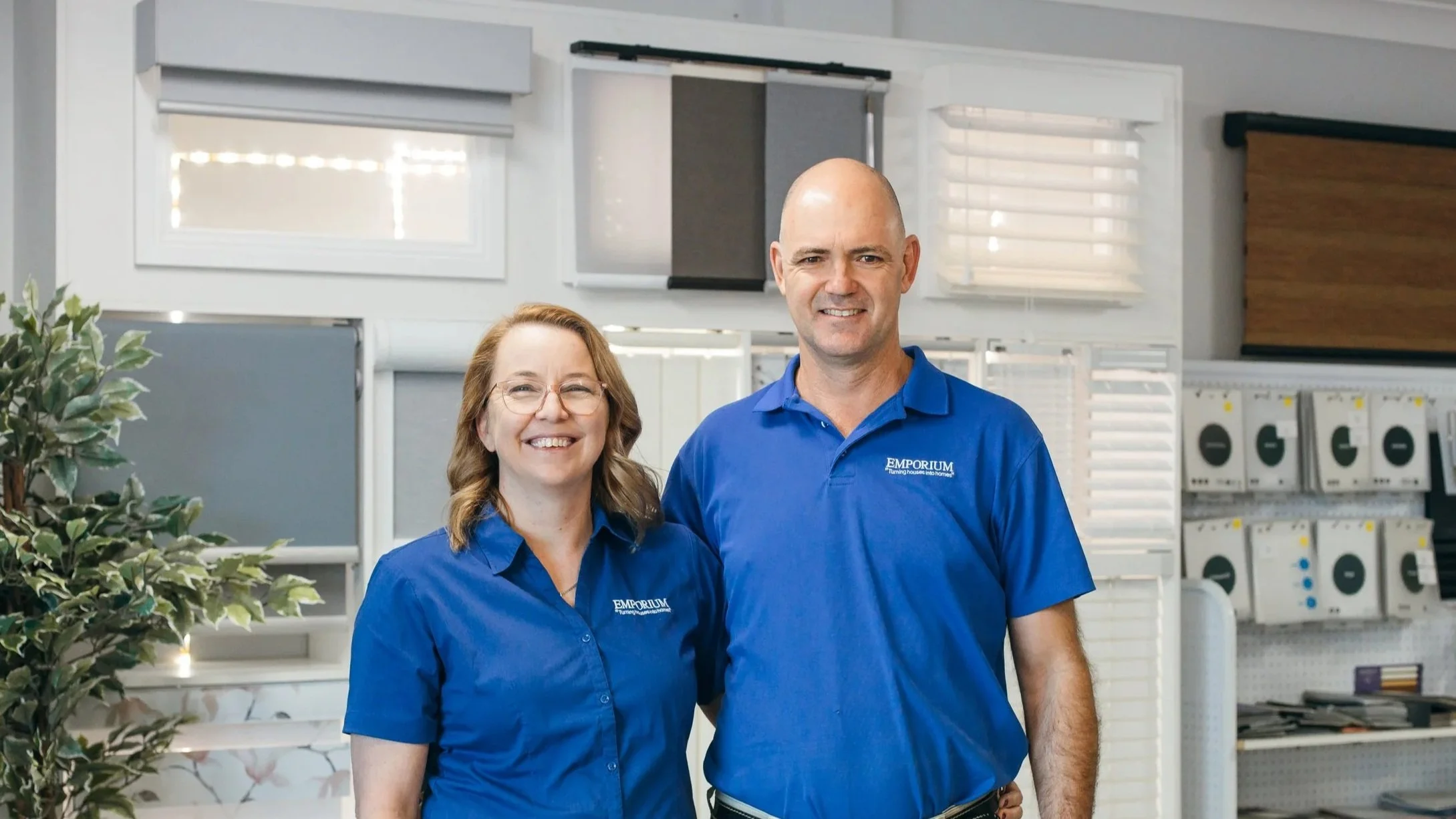 Two employees wearing blue polo shirts with 'EMPORIUM' logo, smiling, standing in front of store equipment, with blinds and a plant in the background.