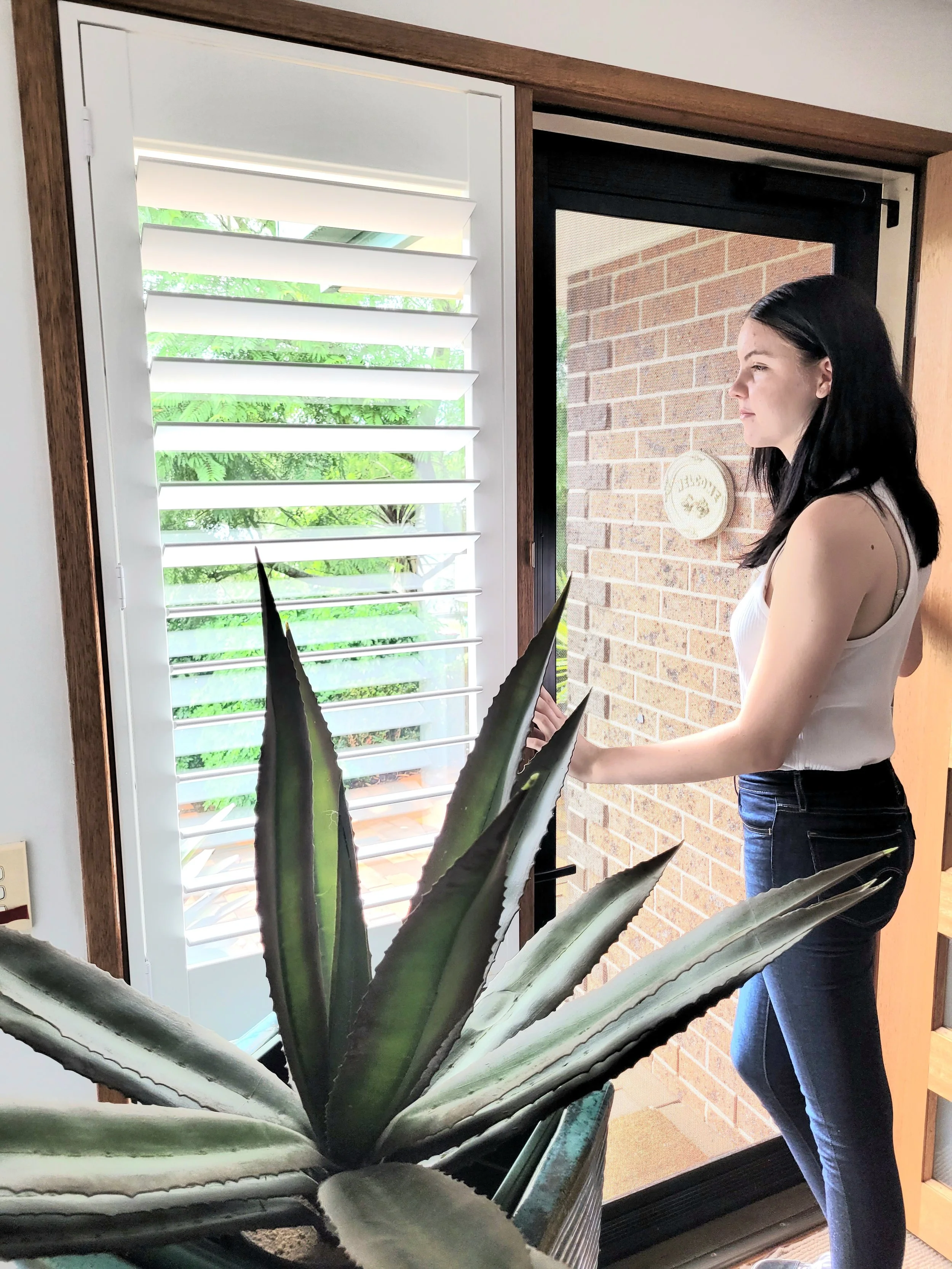 A woman with black hair, wearing a white sleeveless top and blue jeans, stands near a window with white blinds and a brick wall outside. A large green agave plant is in the foreground.