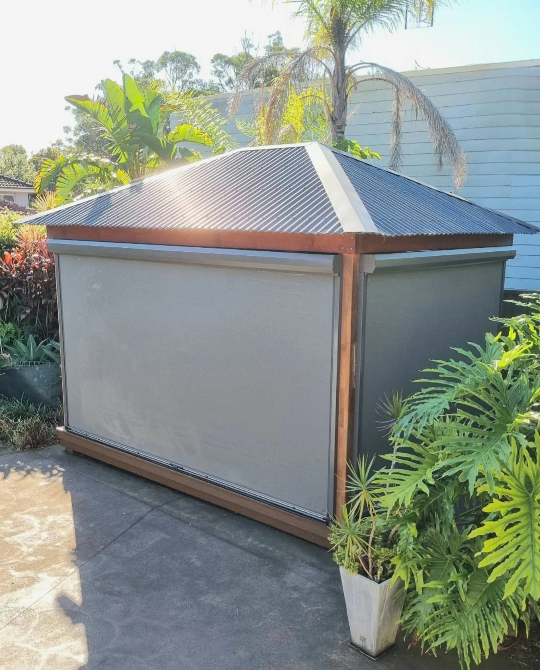 A small outdoor structure with a corrugated metal roof and enclosed sides, placed on a concrete surface, surrounded by lush tropical plants and trees.