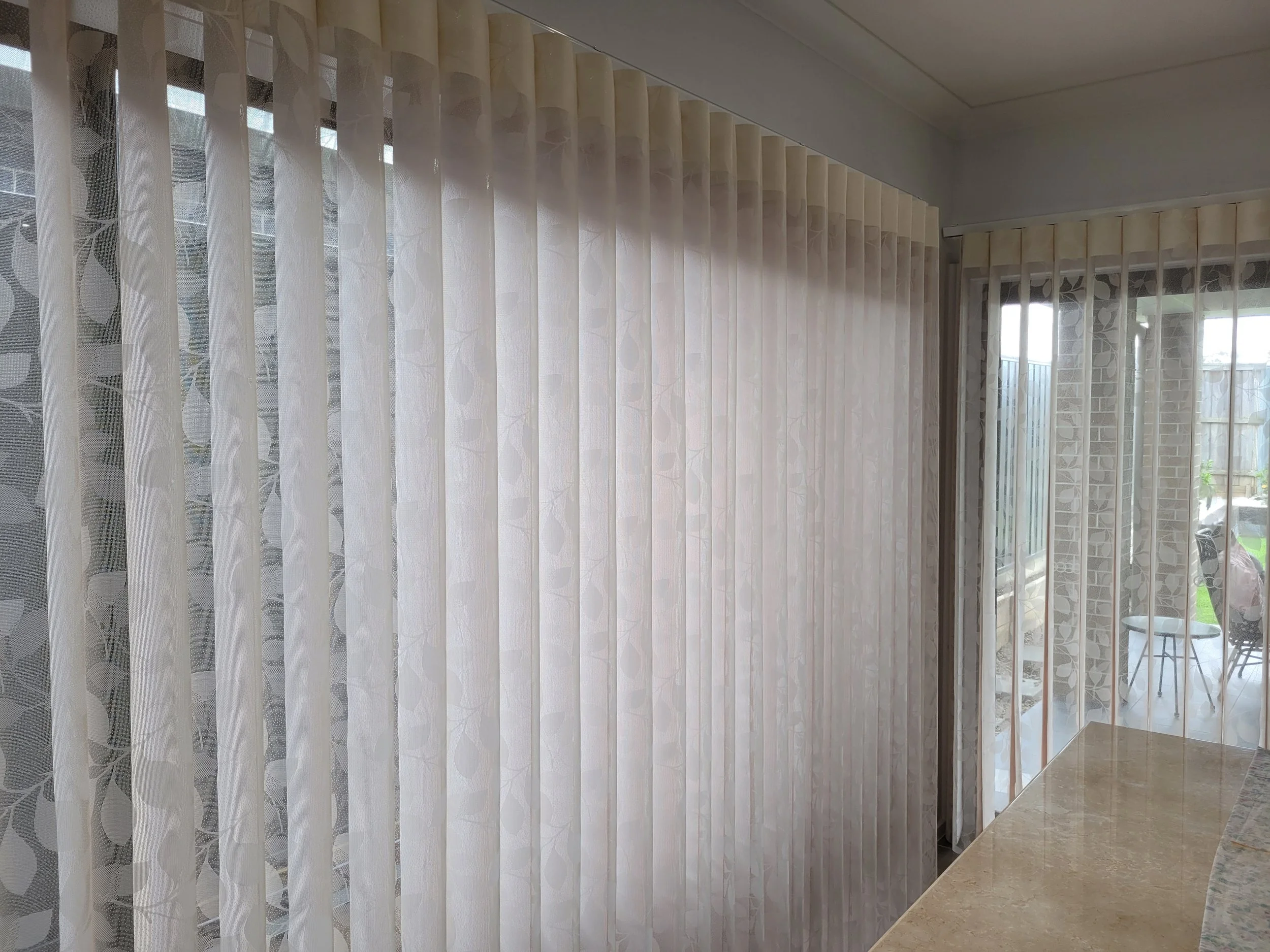 Indoor view of at least two layers of sheer, white patterned curtains covering large windows or sliding glass doors, with a patio and a red brick building visible outside.