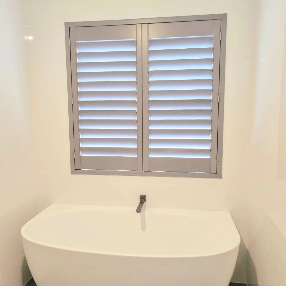 Modern bathroom bathtub below a gray window with white shutters.