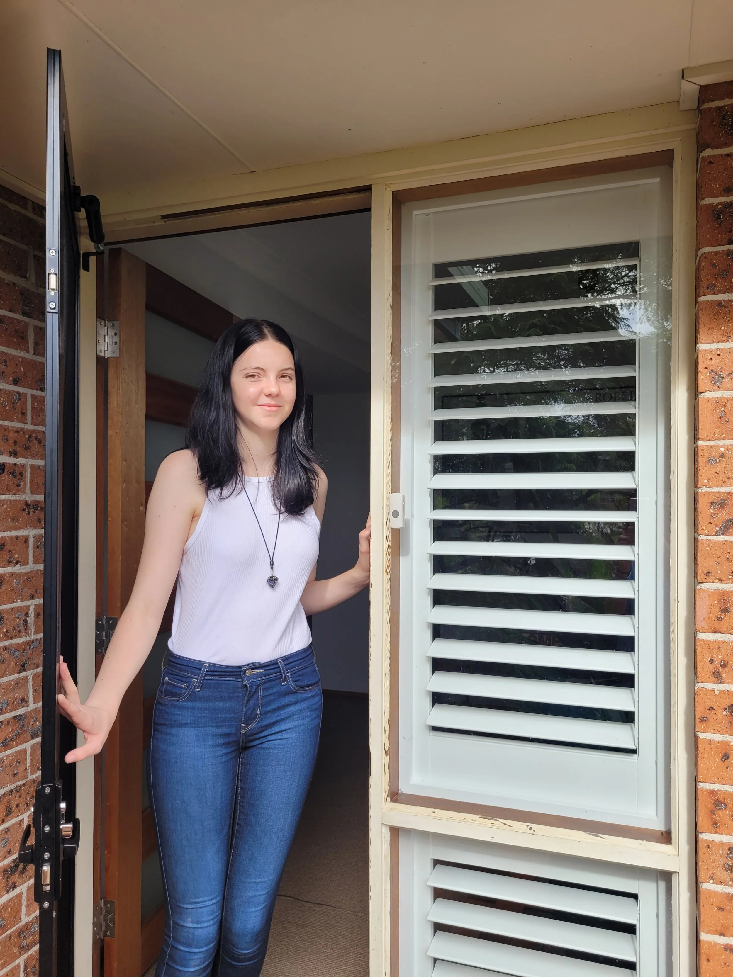 A young woman with long black hair, wearing a white sleeveless top and blue jeans, standing in a doorway. She has a slight smile and holds the doorframe with one hand. Behind her, there is a brick wall, a window with horizontal blinds, and an indoor 