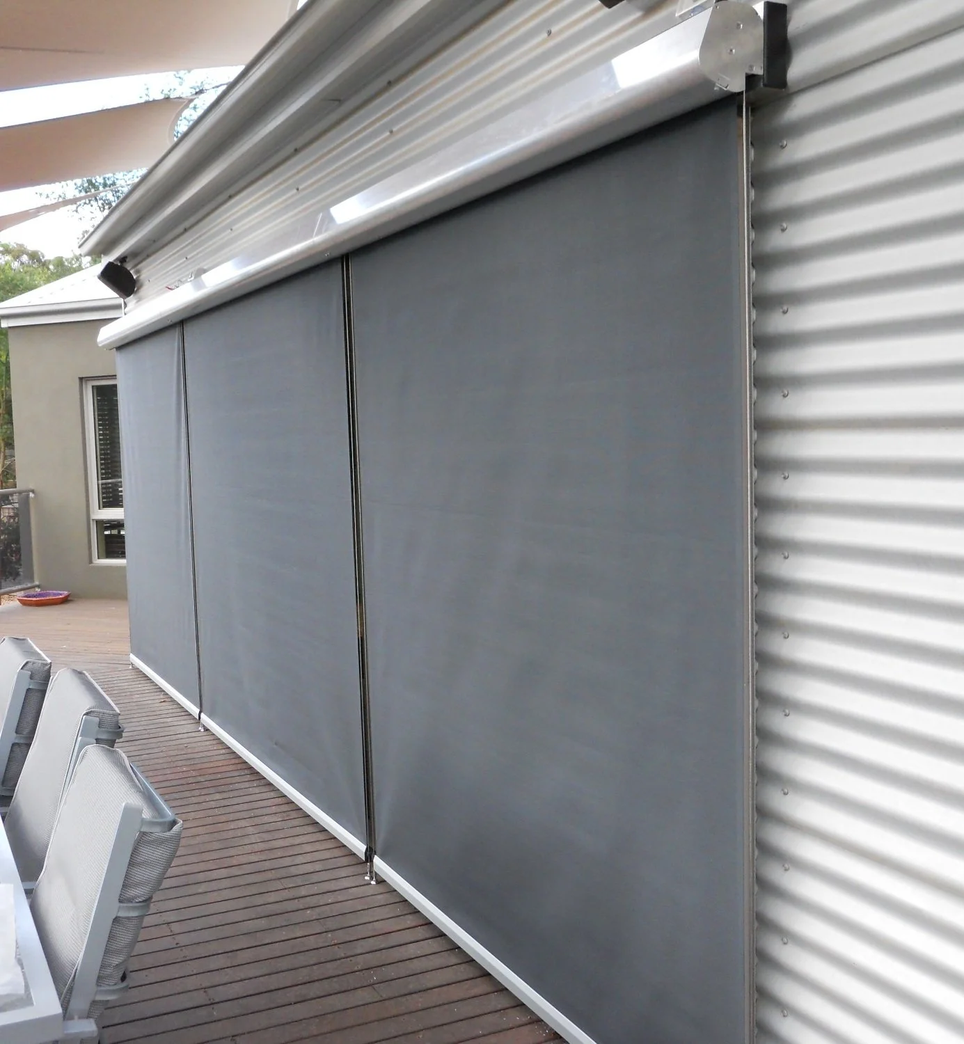 Outdoor patio with a sliding gray roller shade, chairs, and a wooden deck, with a neighboring house visible in the background.
