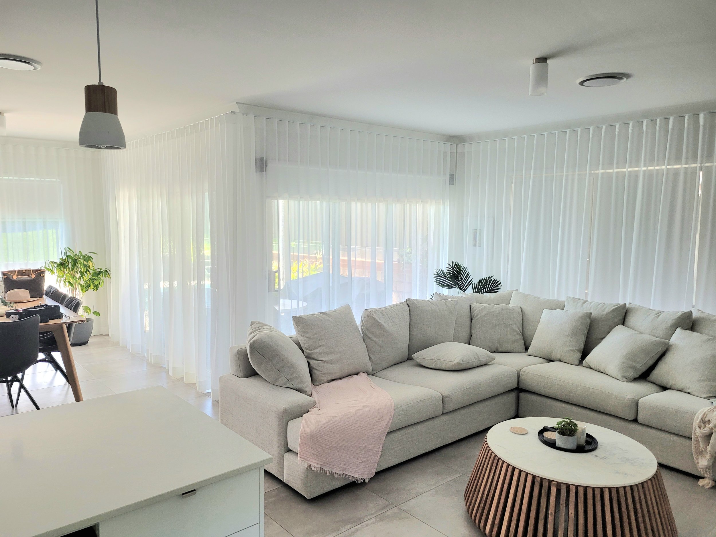 A living room with a large beige sectional sofa, a round wooden coffee table with a white top, and sheer white curtains covering windows. There are some plants and a dining area with black chairs visible in the background. The ceiling has two light f