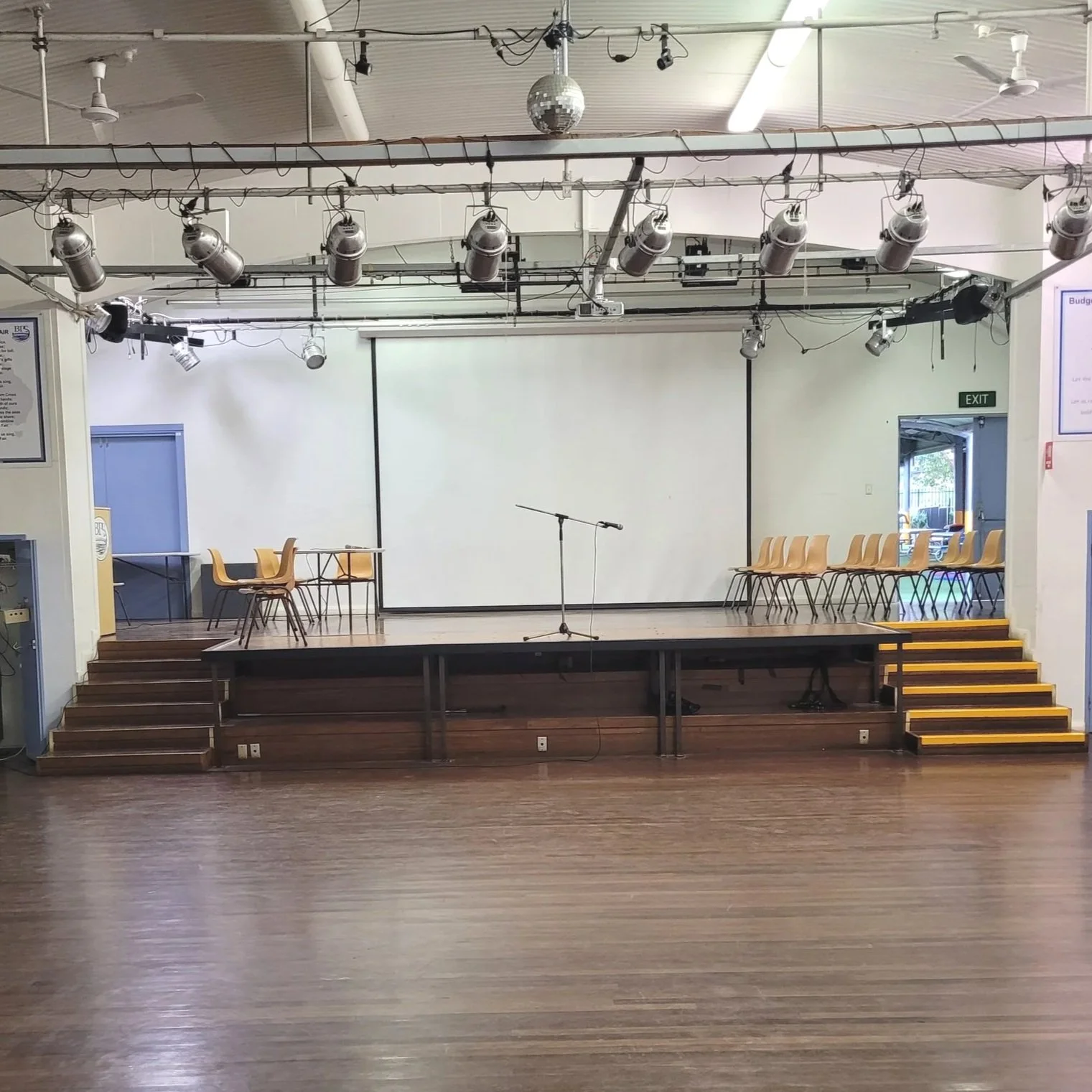Empty stage with a microphone on a stand, surrounded by stacked chairs on each side, in a room with a large blank projection screen and stage lighting above.
