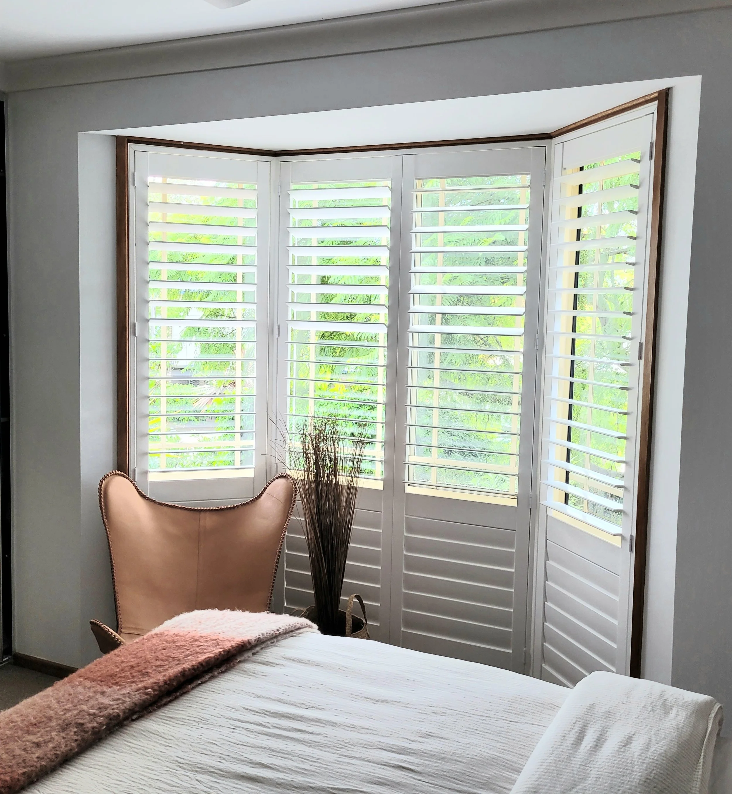 A bedroom with a bed, blanket, and white pillow in the foreground. Behind the bed, there is a window with white plantation shutters, and outside, green foliage is visible. A beige armchair with a pink blanket on top and a tall, dried floral arrangeme