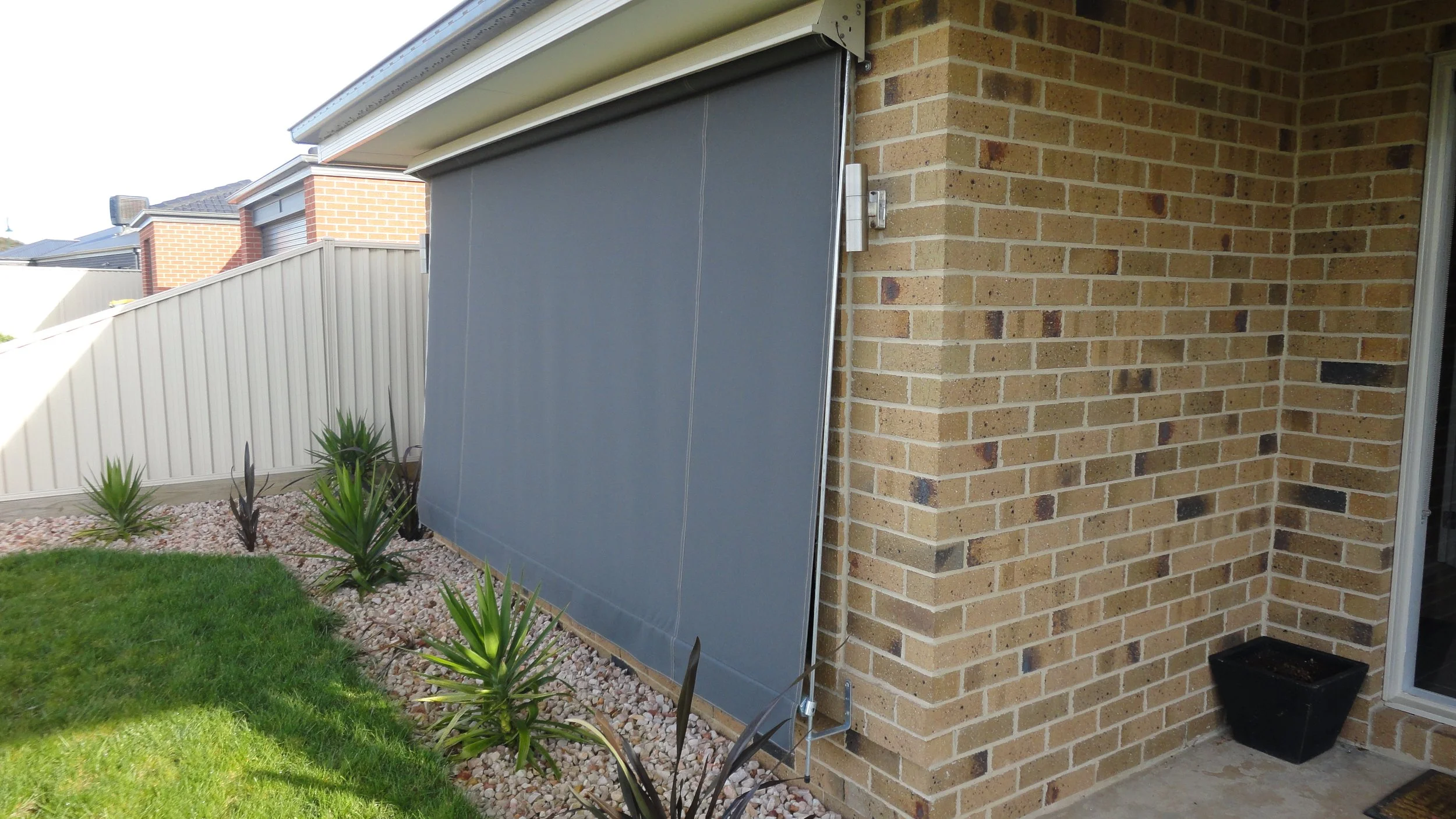 Backyard with brick house wall, grey retractable awning, green grass, small plants, white fence, and patio.