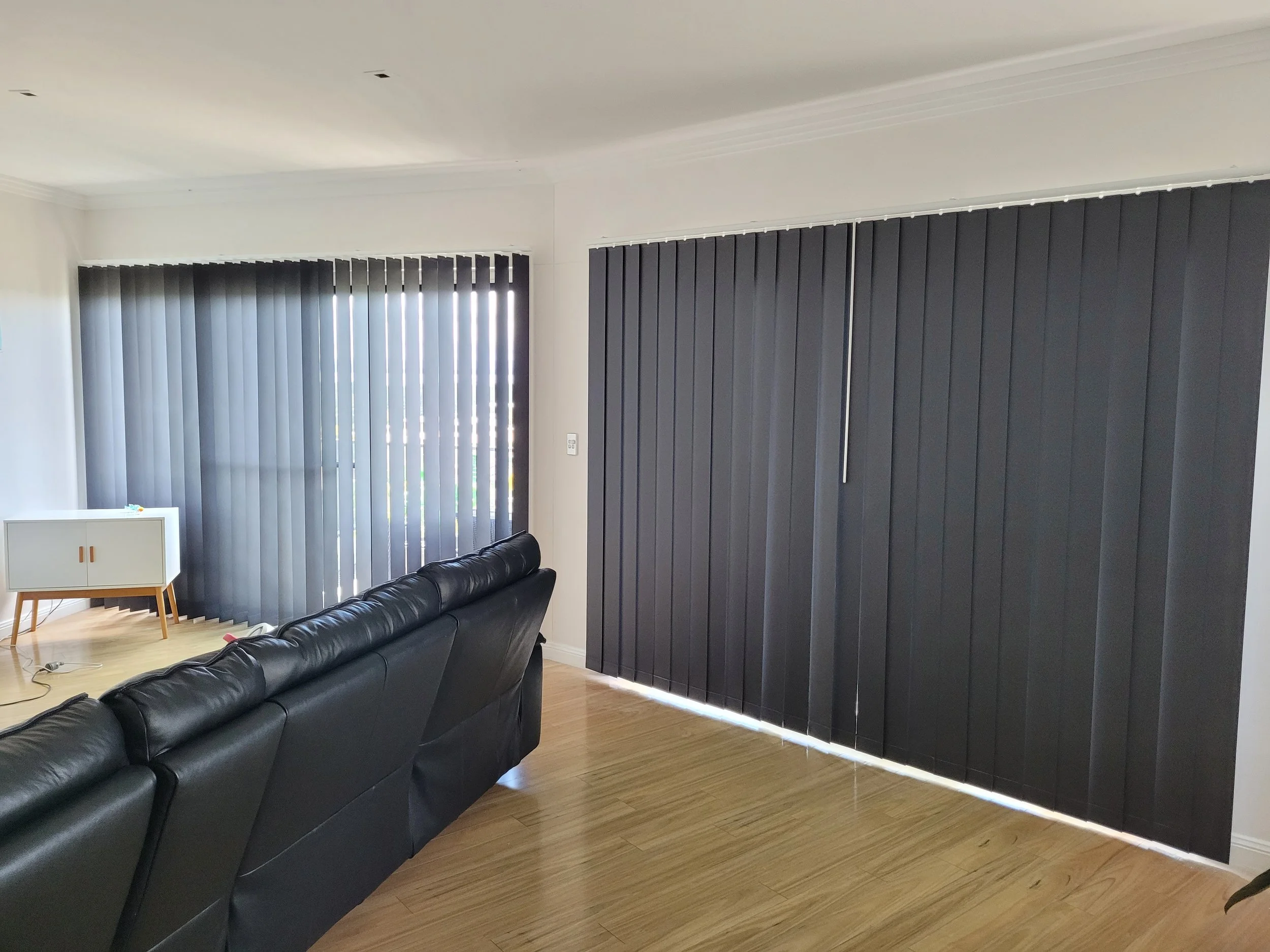 Living room with black leather sofa, white and wood TV stand, and large windows with black vertical blinds.
