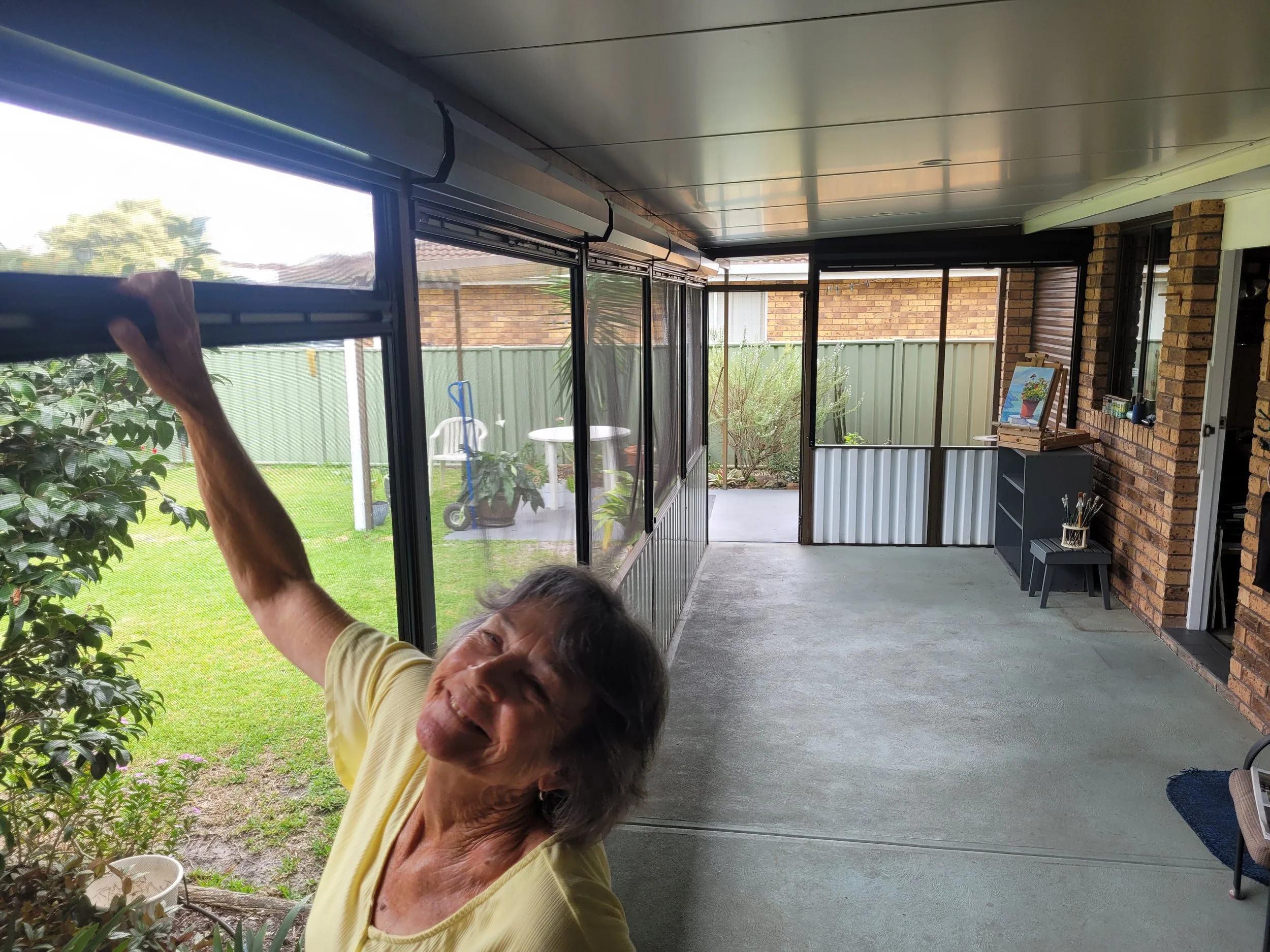 Elderly woman smiling and reaching out with her hand inside a screened porch overlooking a green backyard with trees and a white table and chairs.
