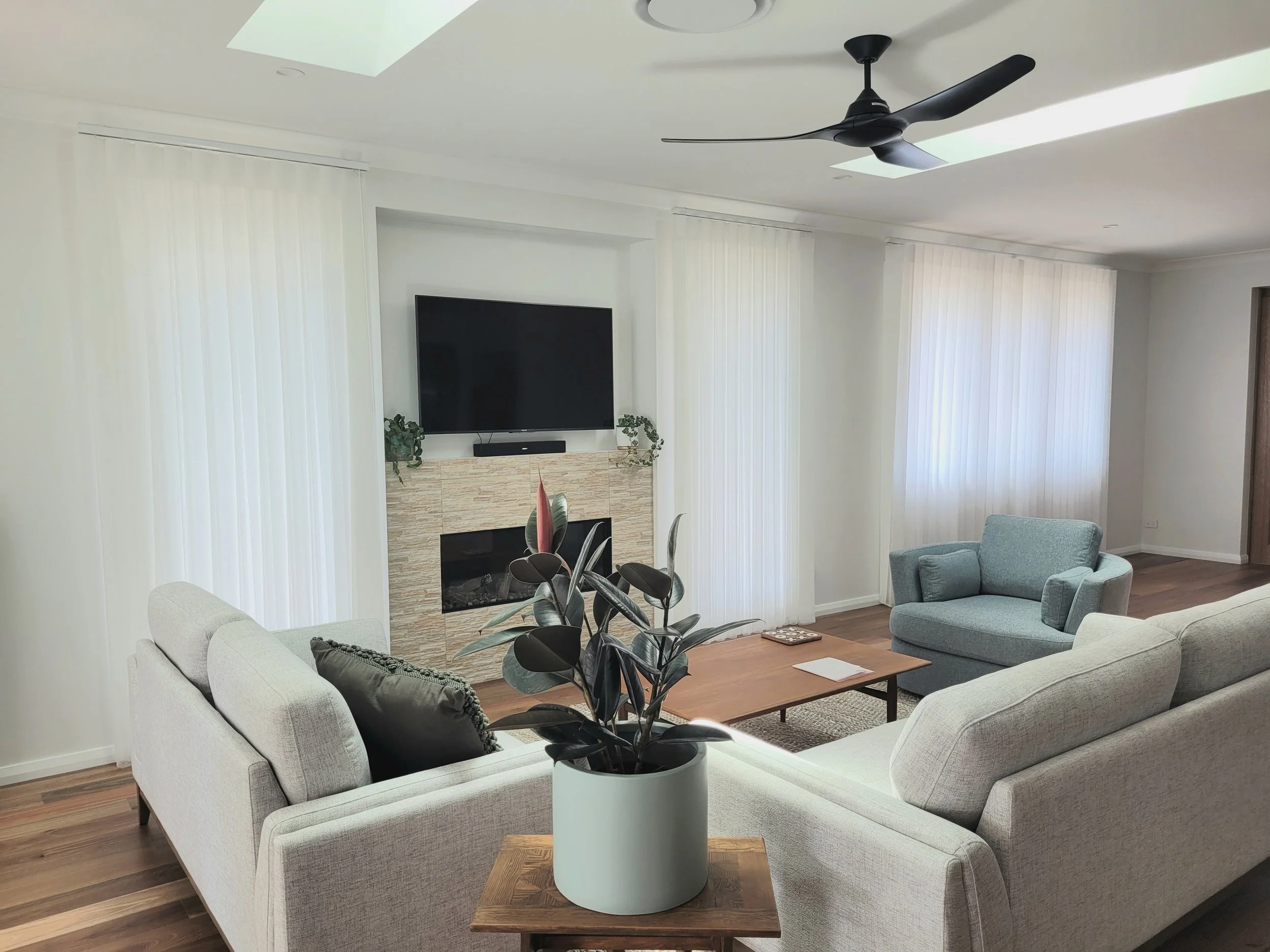 Living room with white upholstered sofas, a wooden coffee table, a potted plant, and a stone fireplace with a mounted flat-screen TV above it. There are vertical blinds covering large windows and a ceiling fan with a light fixture.