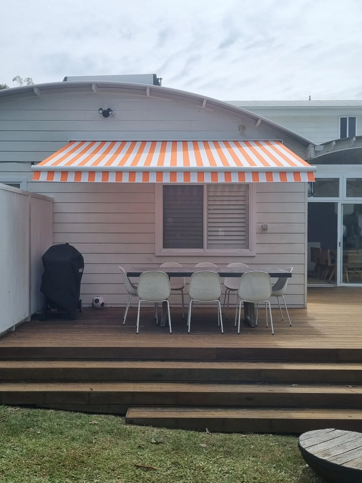 Backyard patio with wooden deck, outdoor dining table and chairs, striped orange and white awning, barbecue grill, soccer ball, and sliding glass doors leading into house.