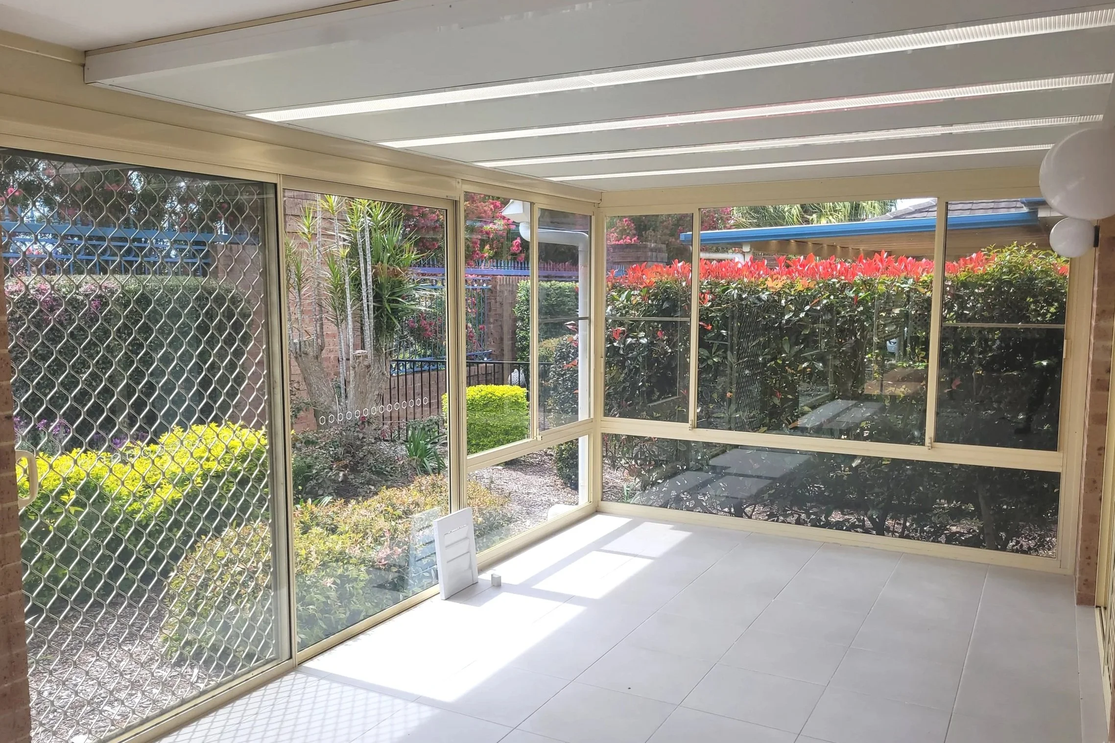 Sunroom with beige-framed glass windows and sliding doors, overlooking a lush garden with green and pink flowering bushes. White balloons are attached to the upper right corner, and the room has white tiled flooring.