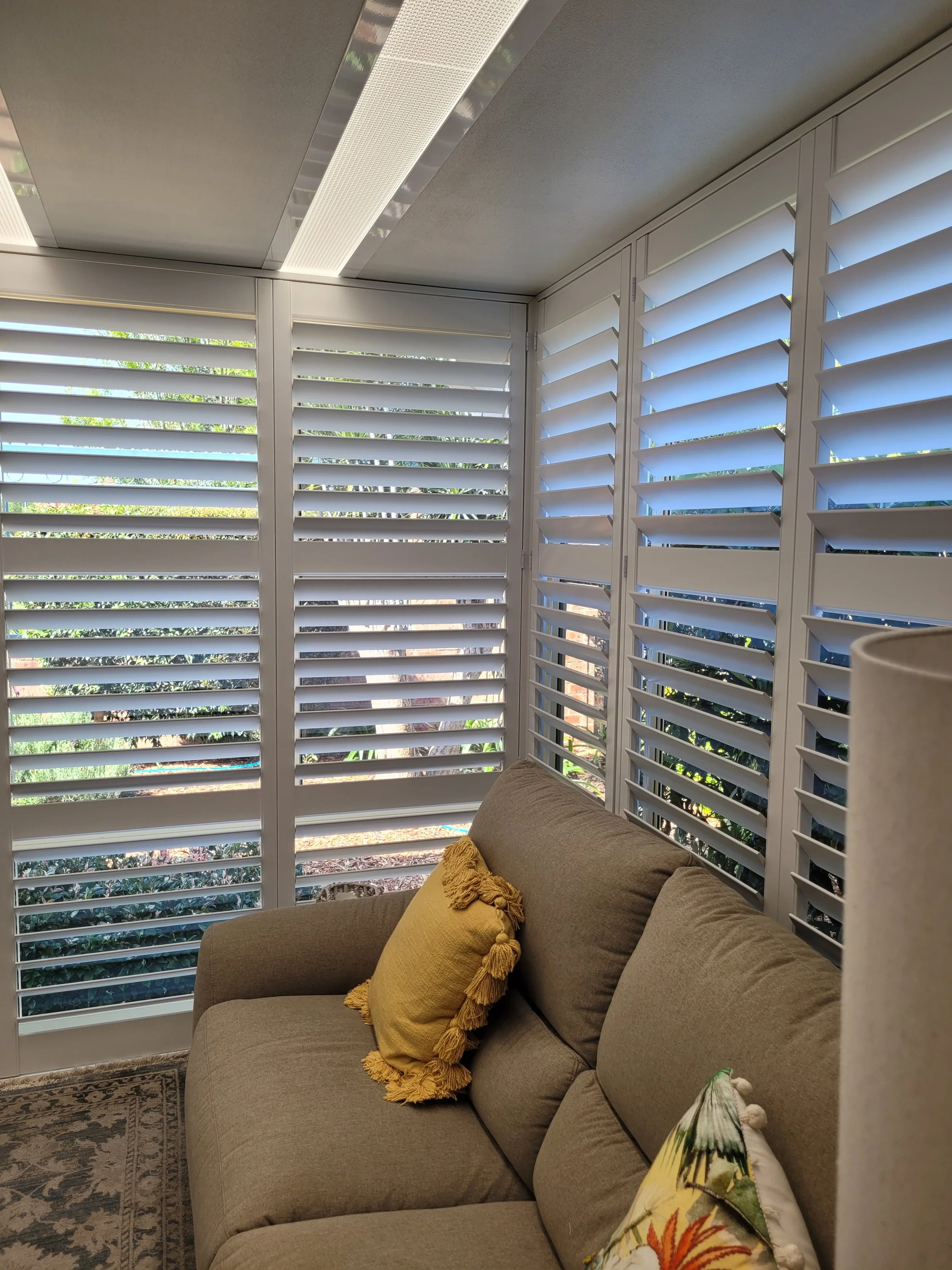 An indoor living space featuring a beige sofa with colorful throw pillows, a patterned rug, and large windows with white plantation shutters that allow sunlight and views of greenery outside.