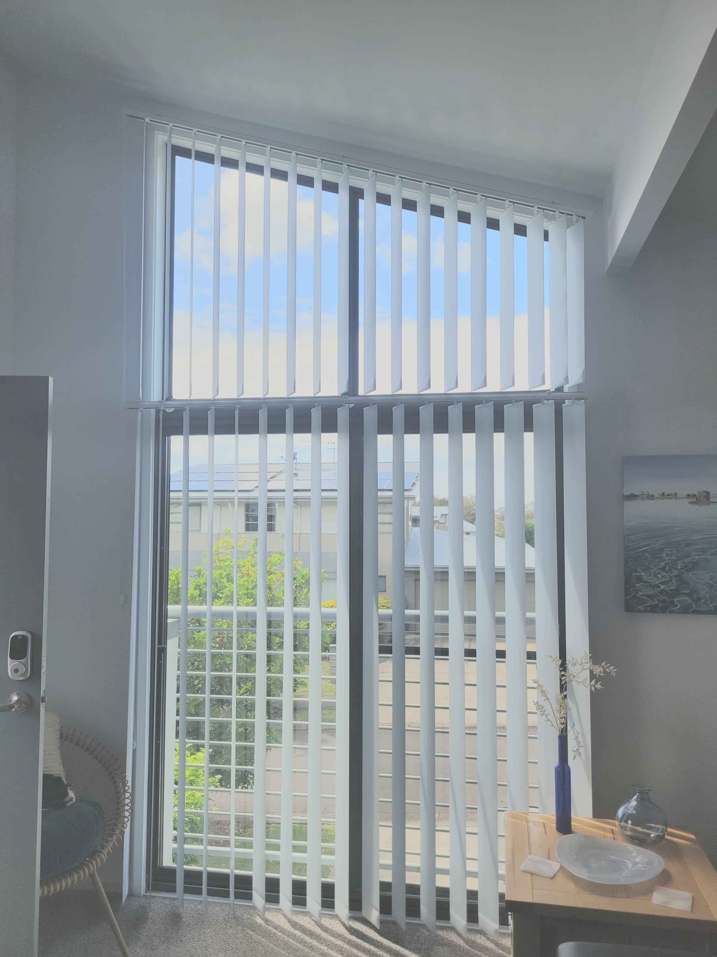 Sunlight coming through vertical white blinds on a sliding glass door, revealing a view of neighboring houses and green trees outside. A small table with a blue vase and decorative items is next to the door.