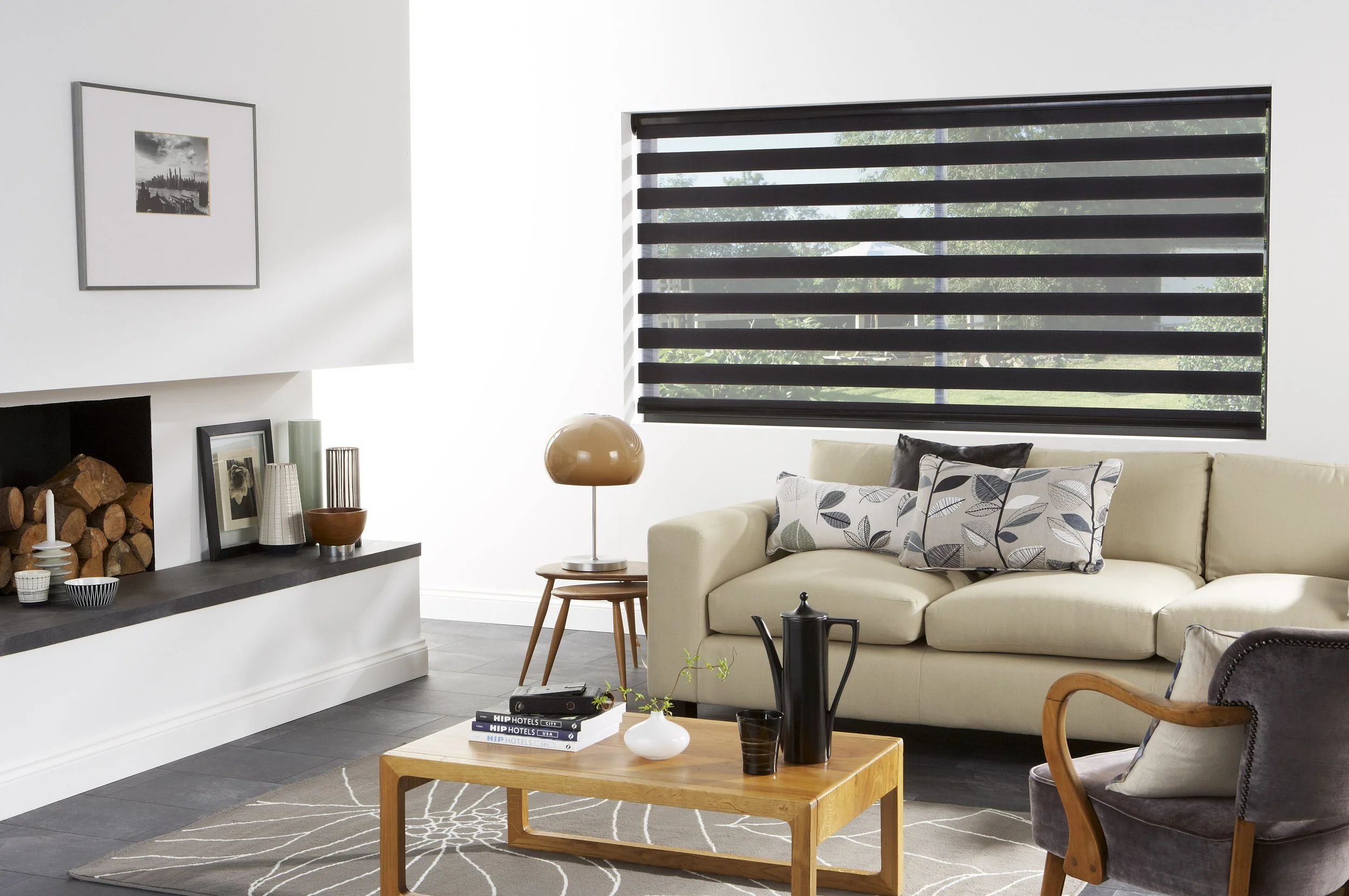 A modern living room with a cream sofa, black and white pillows, a wooden coffee table with magazines and a white vase, a dark purple armchair, a black fireplace with stacked wood, a framed black and white photo on the wall, a brass table lamp, and a