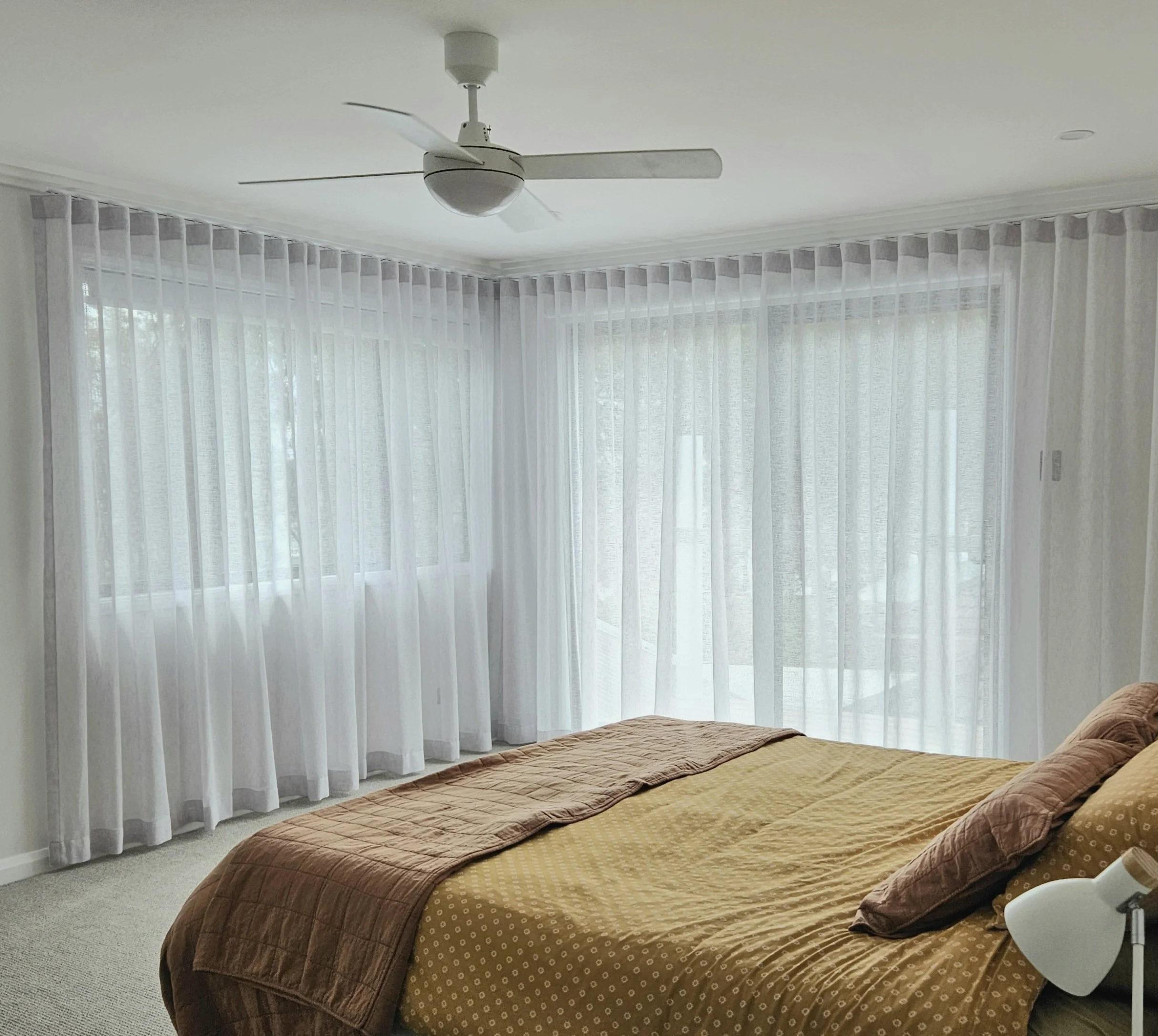 A bedroom with a bed featuring a yellow and brown bedspread, situated near a large window with sheer white curtains, a ceiling fan, and a bedside lamp.