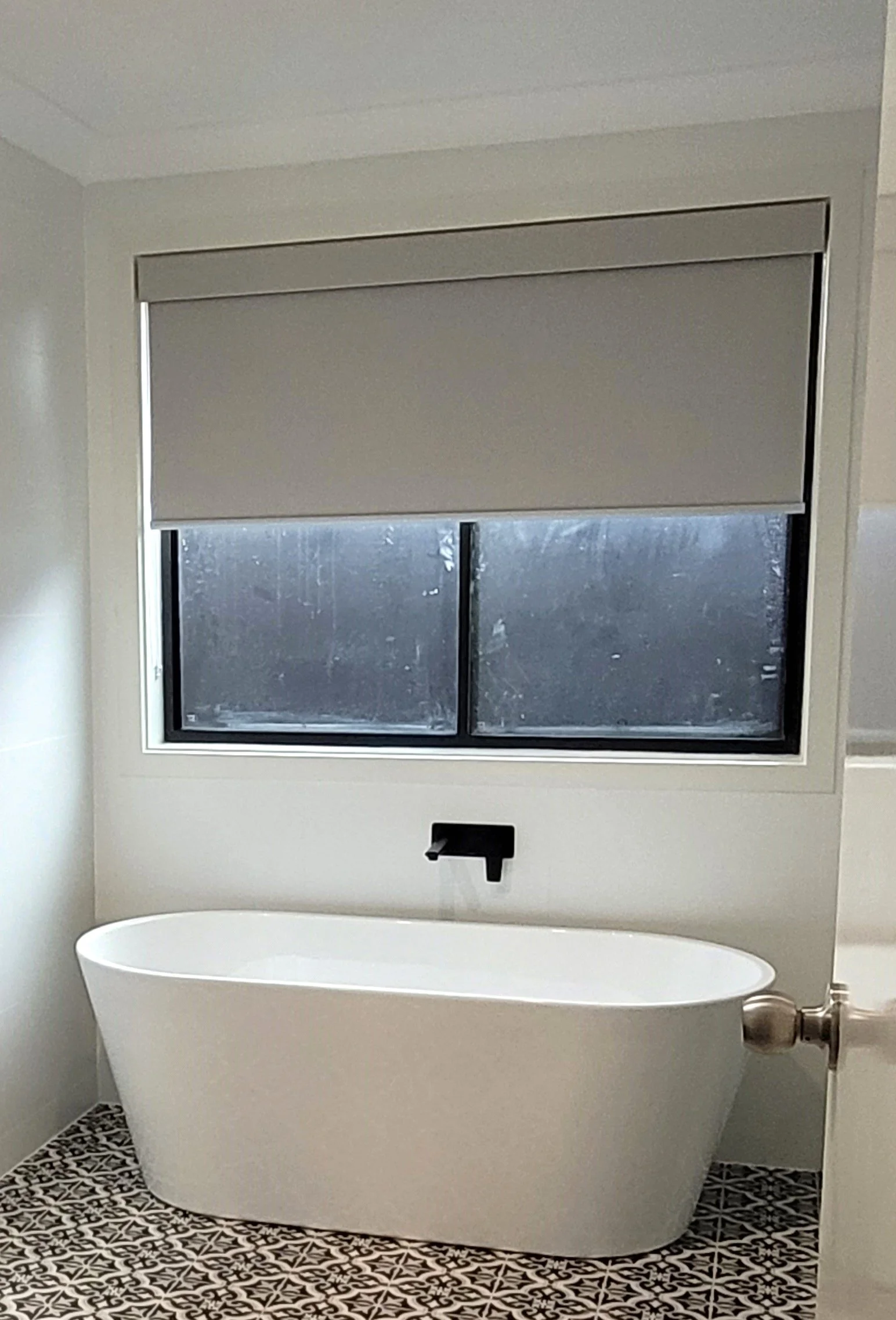 A bathroom with a white bathtub, a window with a gray roller shade, and patterned floor tiles.