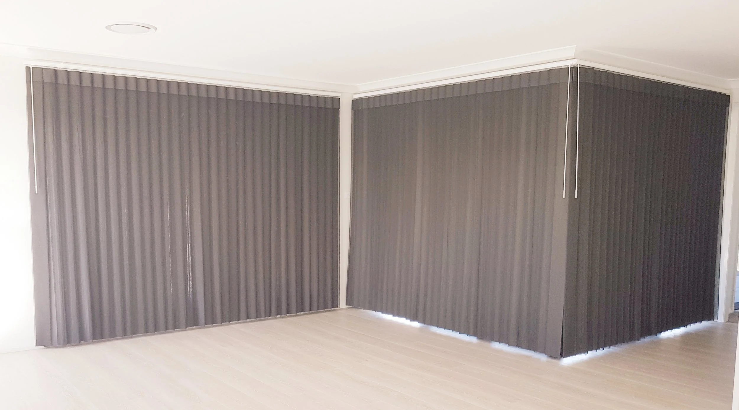 Gray vertical blinds on a window, partially closed, in a room with light-colored wooden flooring and white walls.