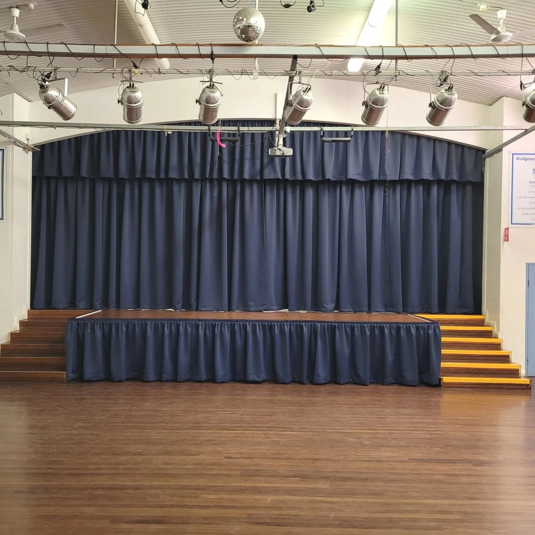 Empty stage with blue curtains, wooden steps, and a disco ball hanging from a metal rail with spotlights in a large hall or auditorium.