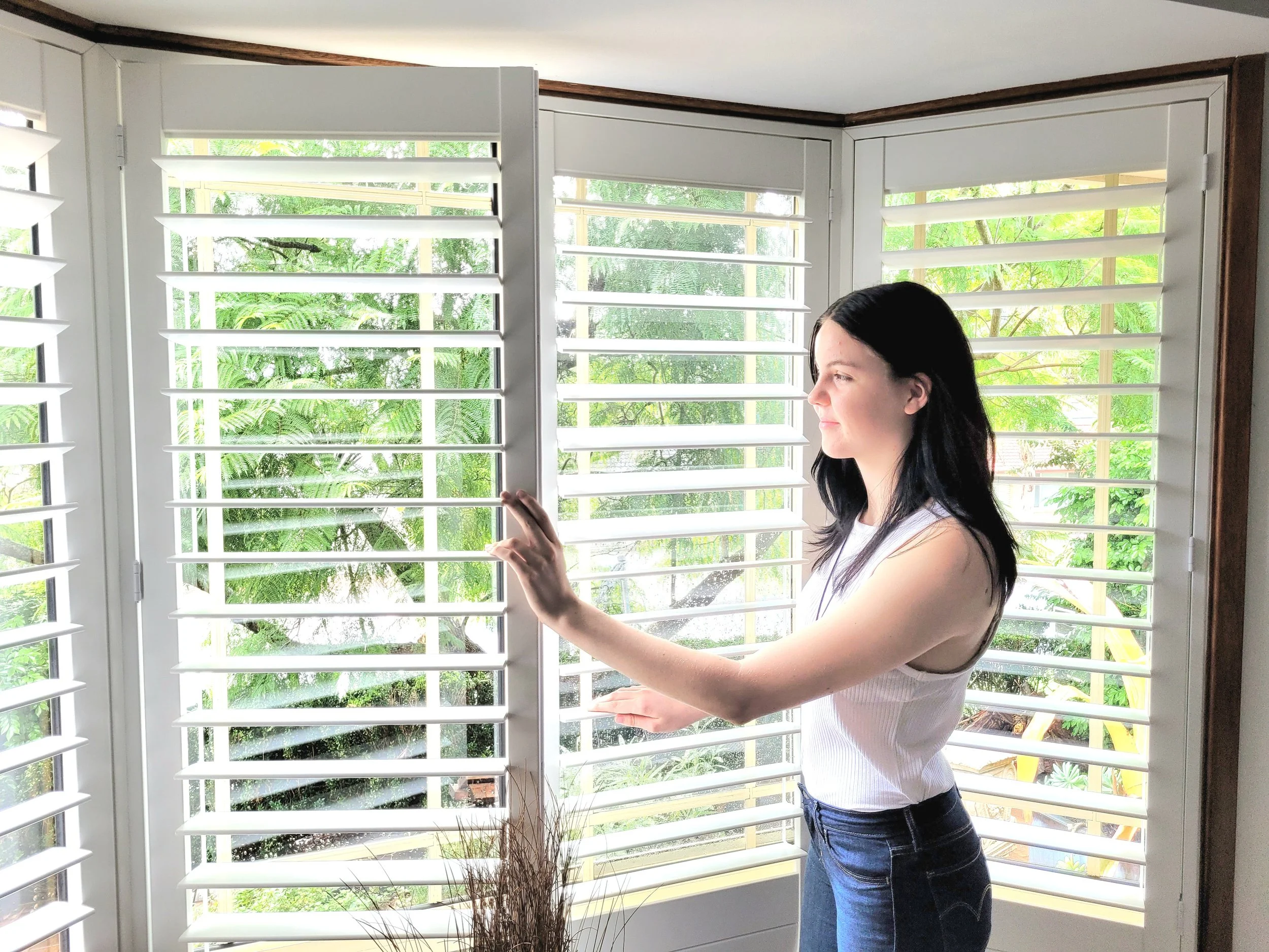 A woman with black hair wearing a white sleeveless top and blue jeans, standing by white window shutters, holding one shutter open to look outside at lush green trees.
