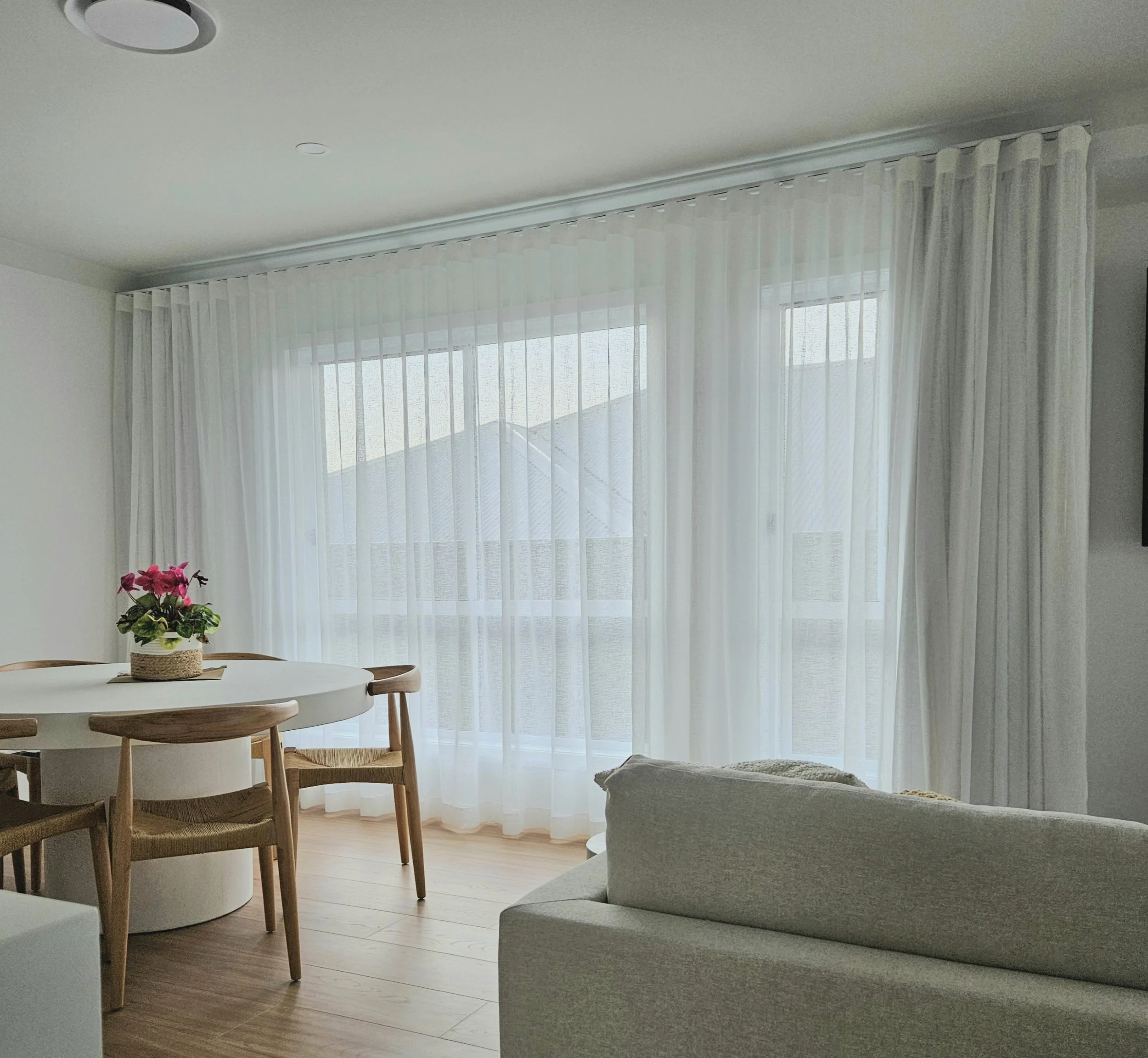 Living room with sheer white curtains, a round white dining table with a potted pink flowering plant, wooden chairs, and a beige upholstered sofa, with a ceiling light fixture.