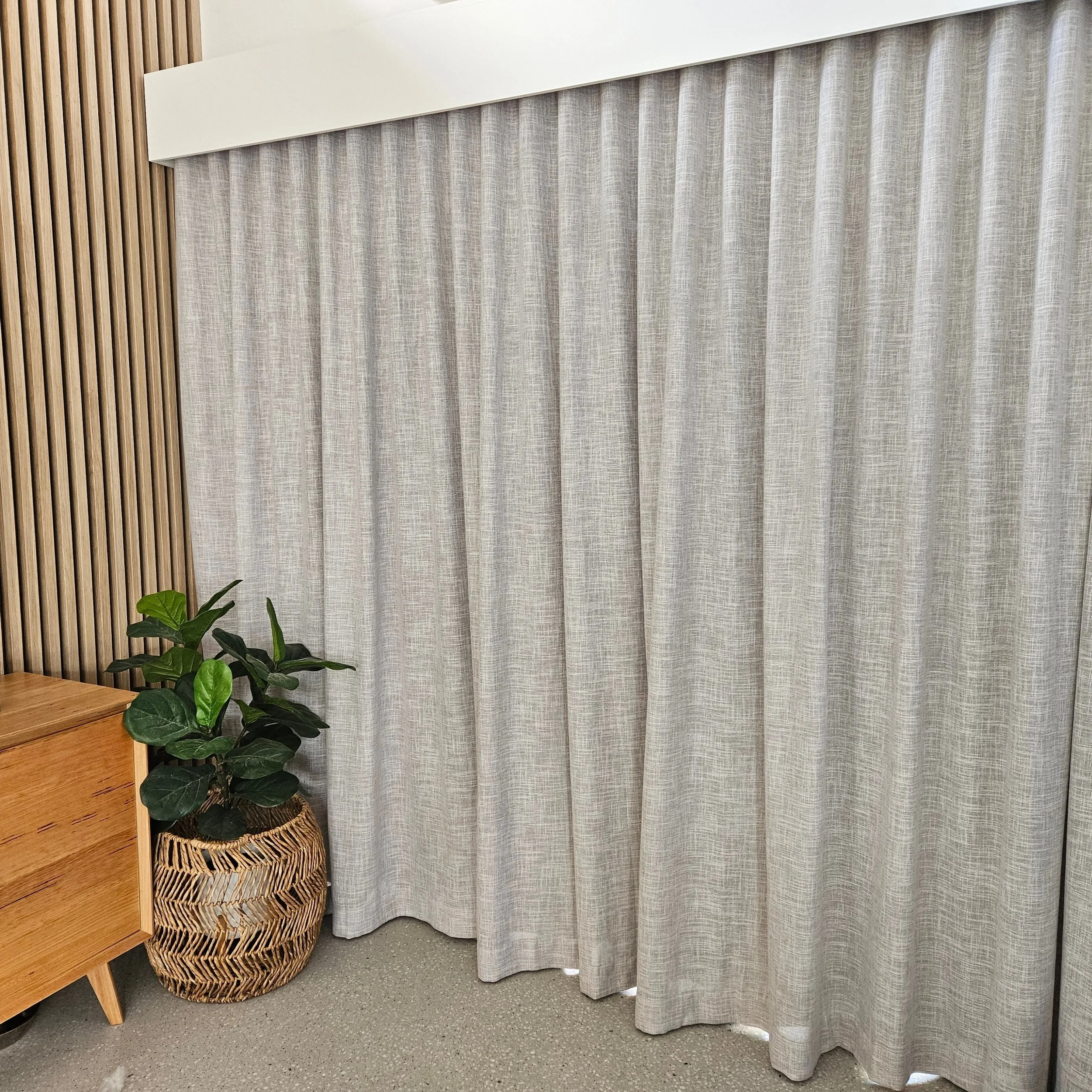A room corner featuring a beige curtain, a wooden slat wall, a wooden cabinet, a woven basket, and a green potted plant on the floor.