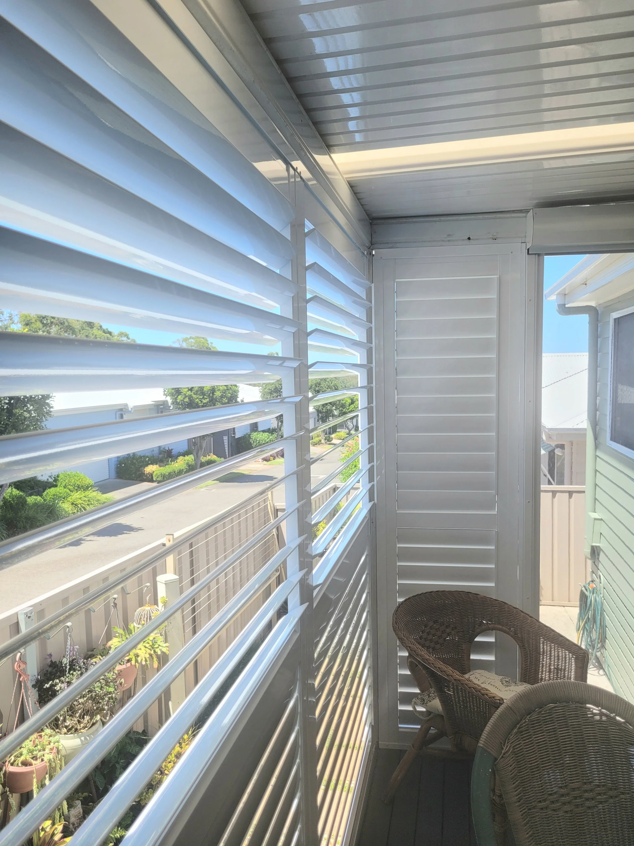 A small covered balcony with a wicker chair, white horizontal window shutters, and a view of flowers and trees outside.