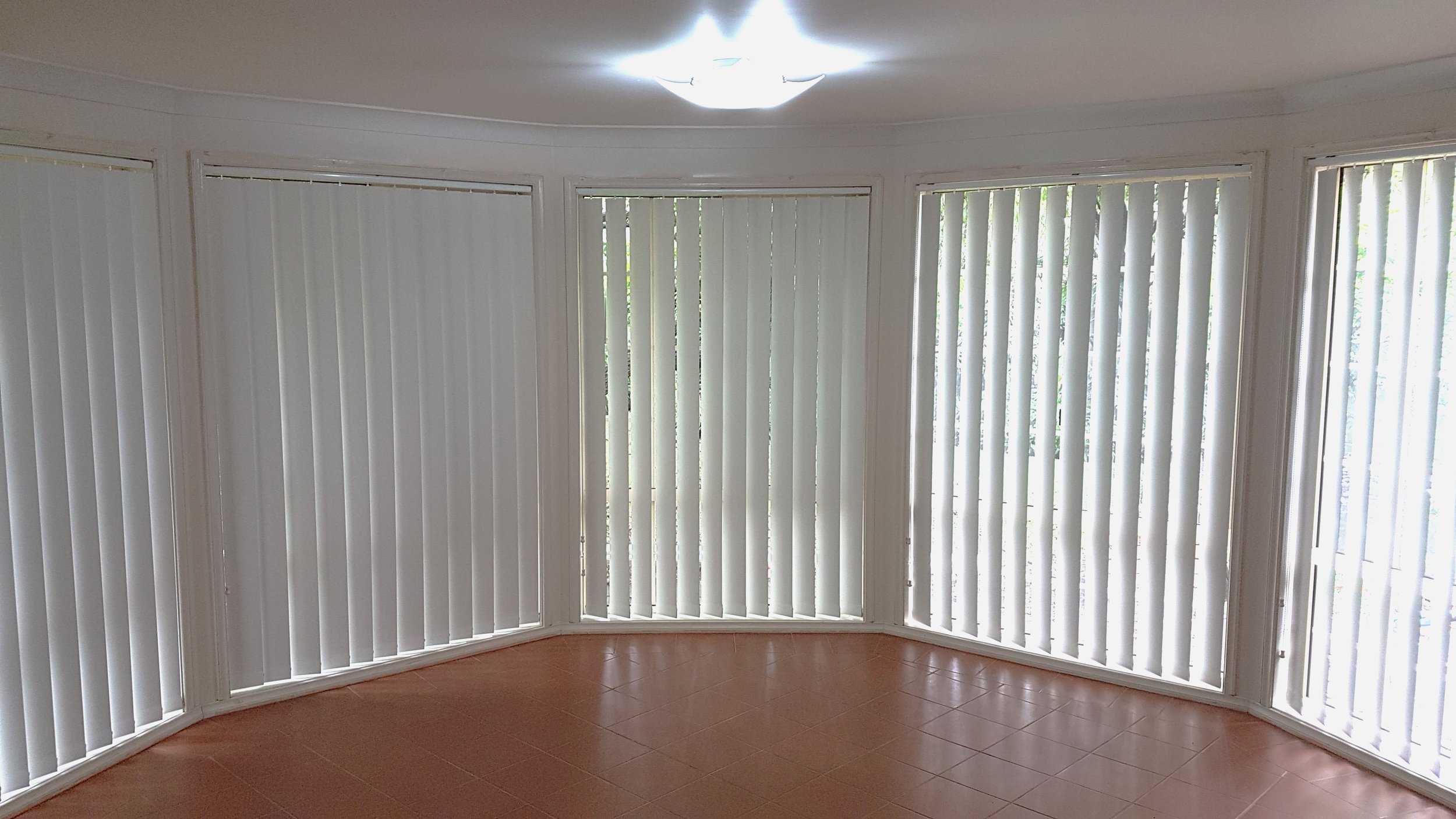 Living room with large bay windows covered by white vertical blinds, tiled floor, and a ceiling light fixture.