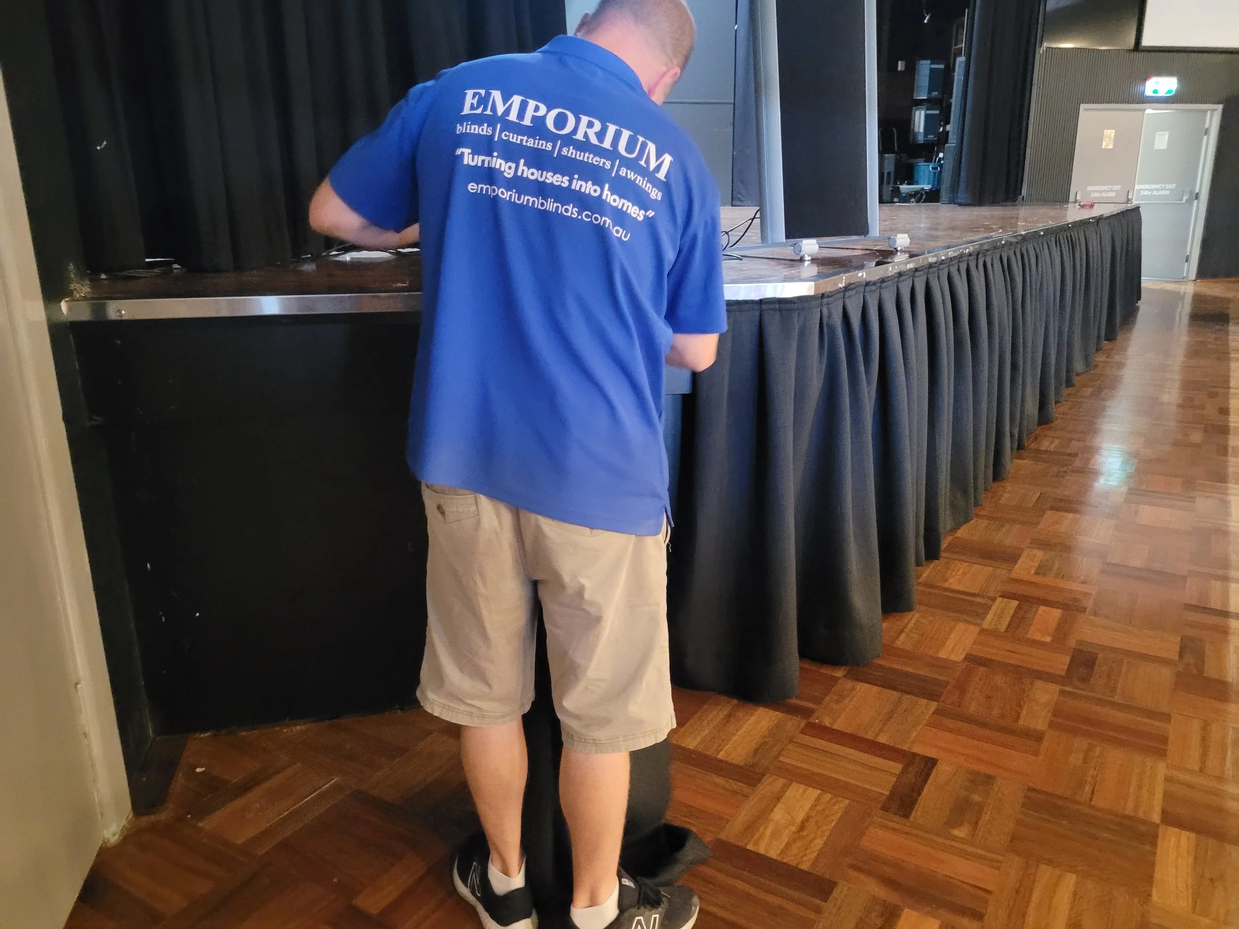 A person wearing a blue shirt with the logo 'EMPORIUM' on the back is helping another person put on shoes in an indoor hall with a stage and exit doors in the background.