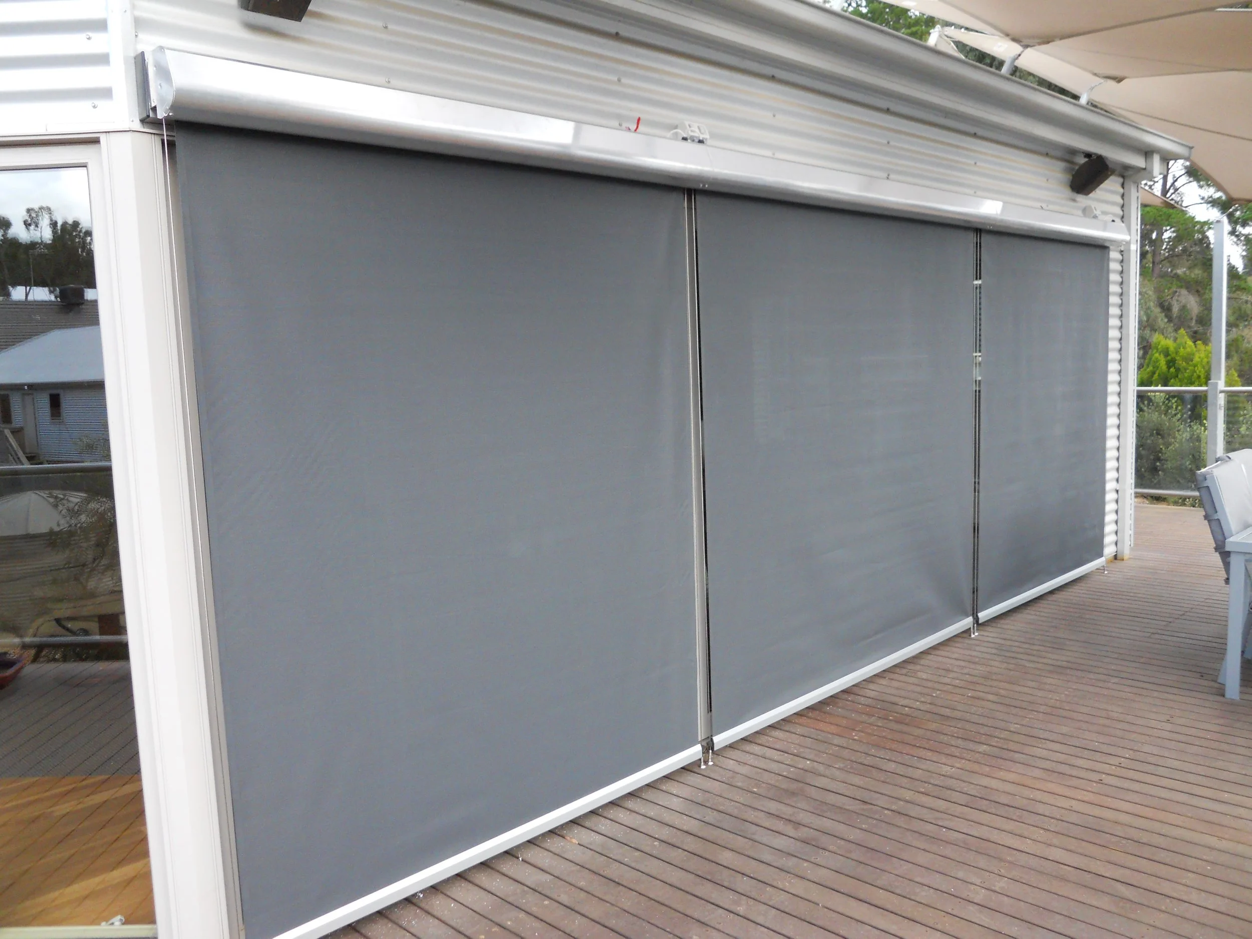 An outdoor balcony with a wooden floor, large sliding glass doors, and a gray retractable screen or curtain. There is a white railing with trees and rooftops in the background.