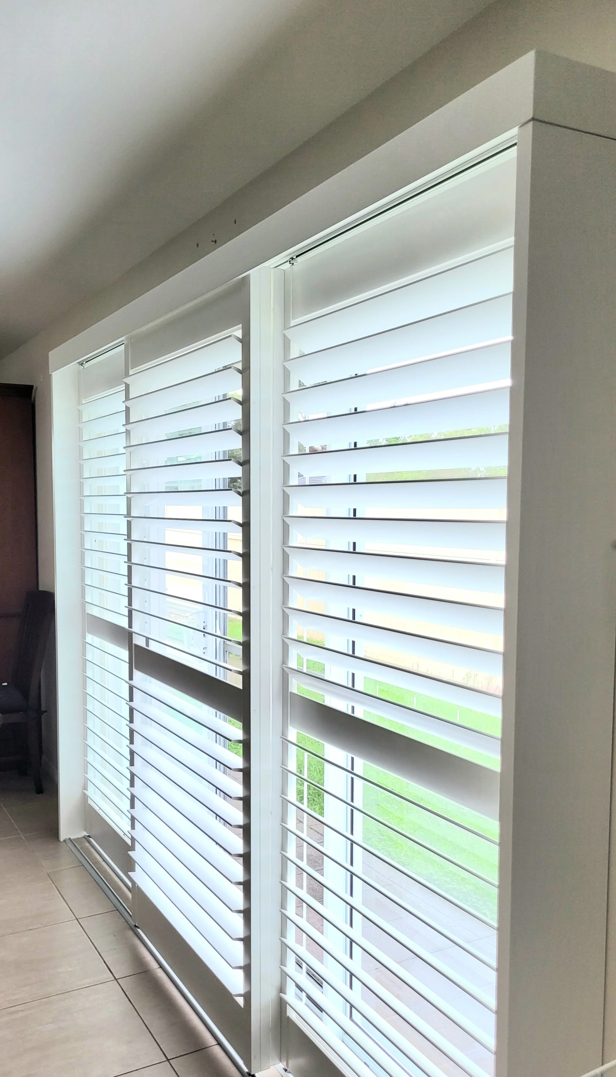 Interior view of white plantation shutters covering large windows, with a glimpse of green grass outside. The room has tiled flooring and part of a dark chair is visible on the left.