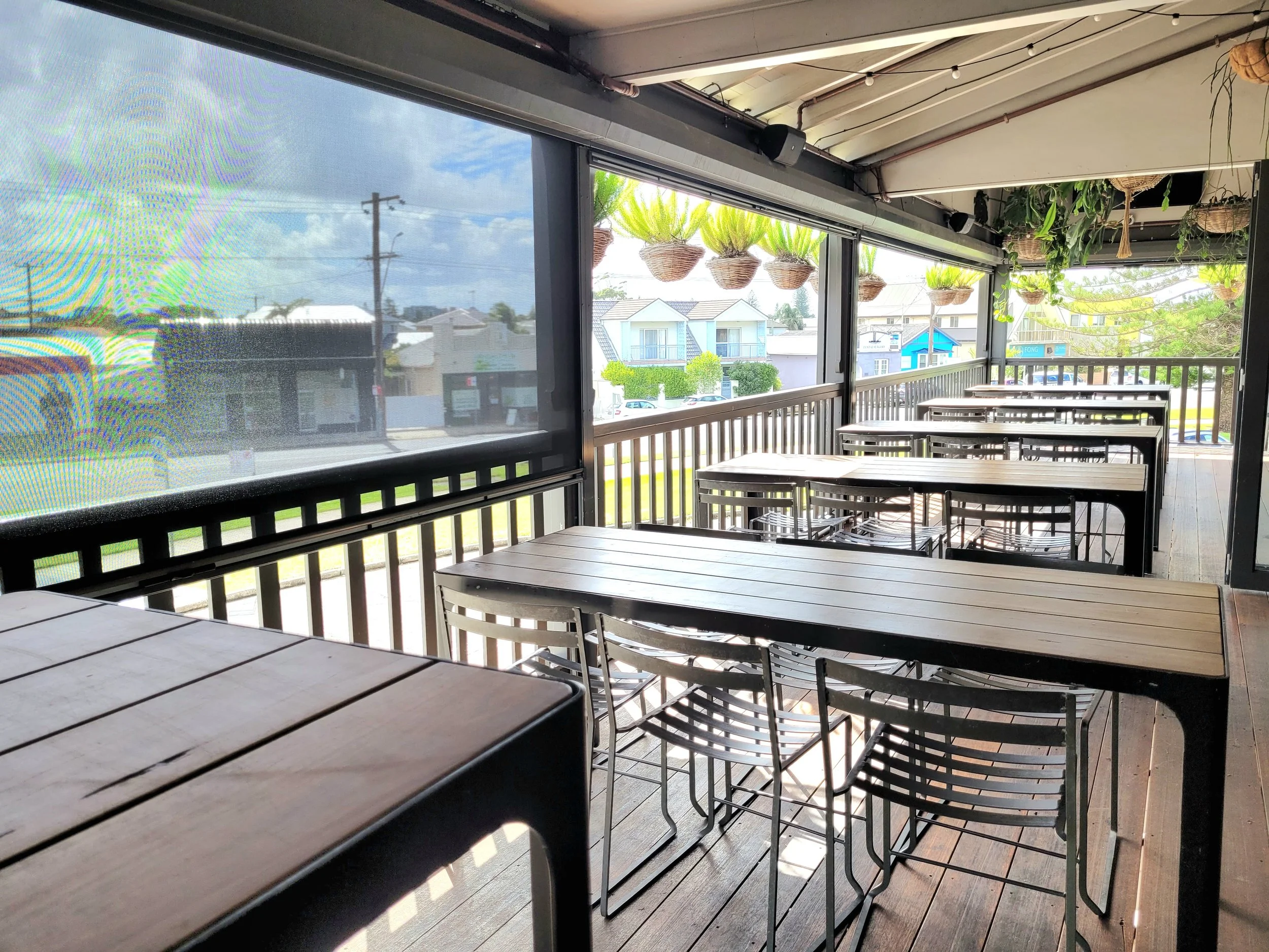 Outdoor patio with wooden tables and metal chairs, hanging plants in woven baskets, and a screened window showing a cloudy sky and nearby houses.