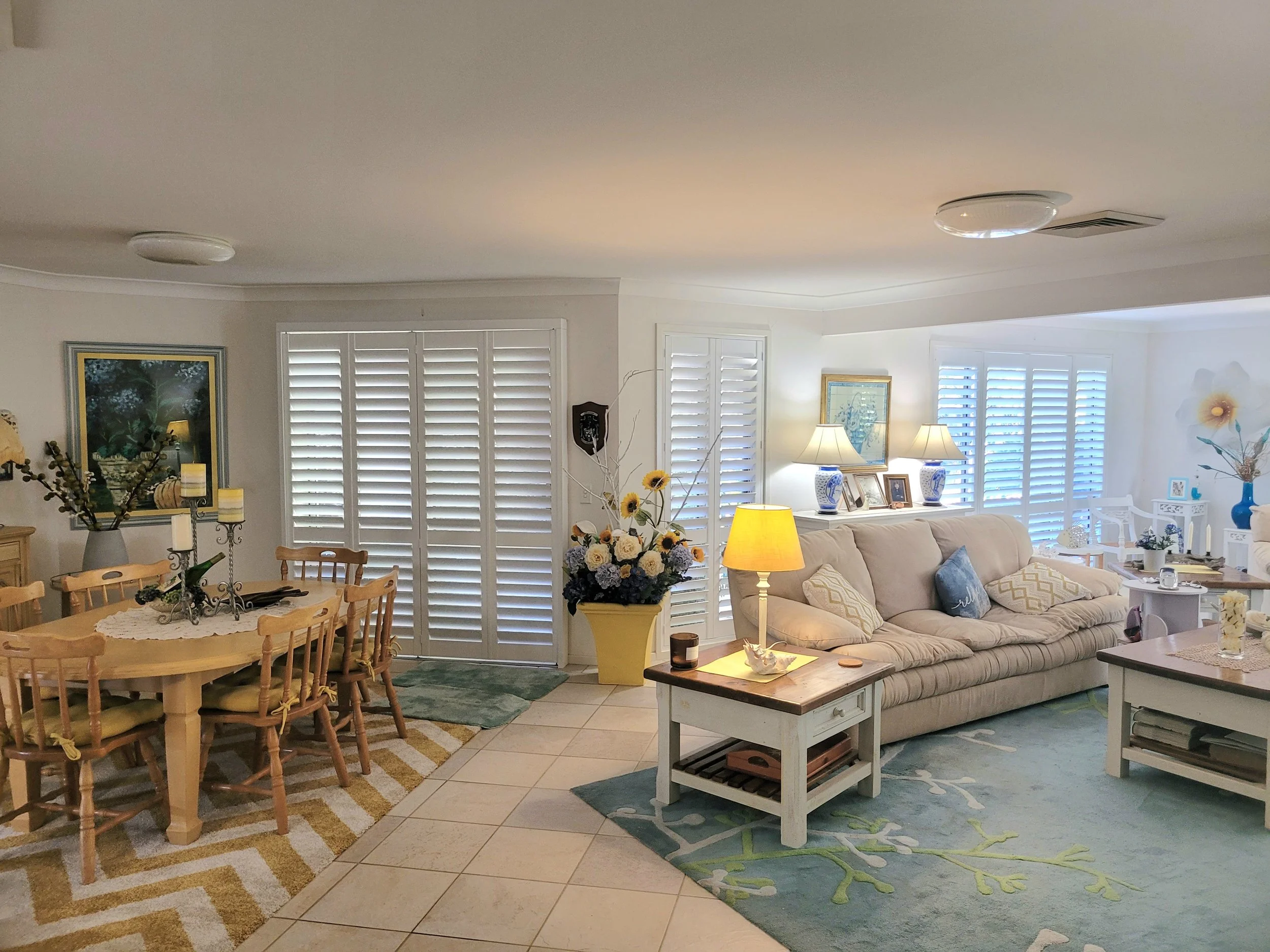 Interior of a living room and dining area with white shutters on the windows, a beige sofa with pillows, a wooden dining table with chairs, floral arrangements, lamps, and decorative items, and a multicolored area rug.