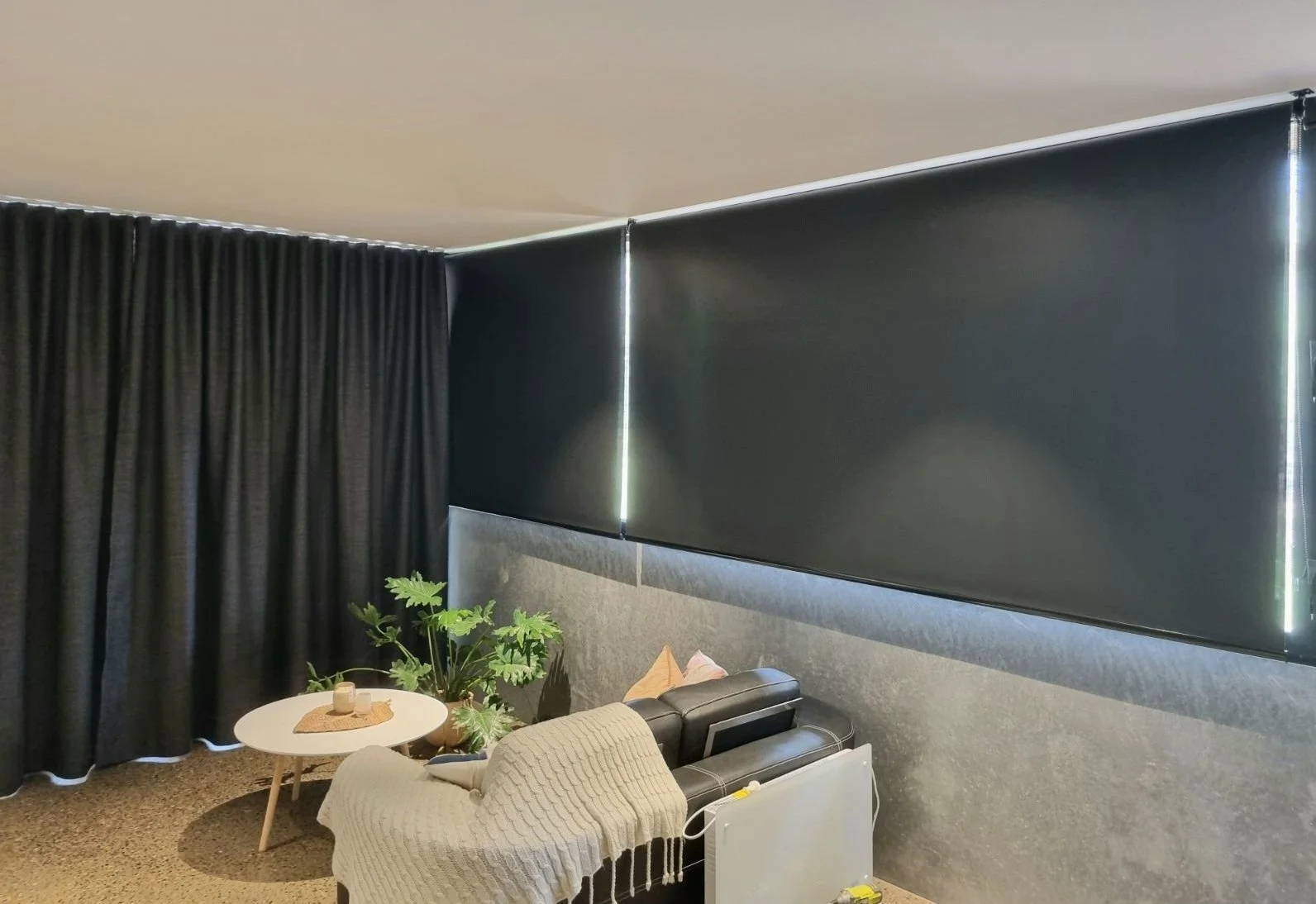 Living room with black curtains, black window shades, a gray upholstered headboard, a small white round table with a plant and candles, a beige throw blanket on a white sofa with pillows, and a black leather armchair.
