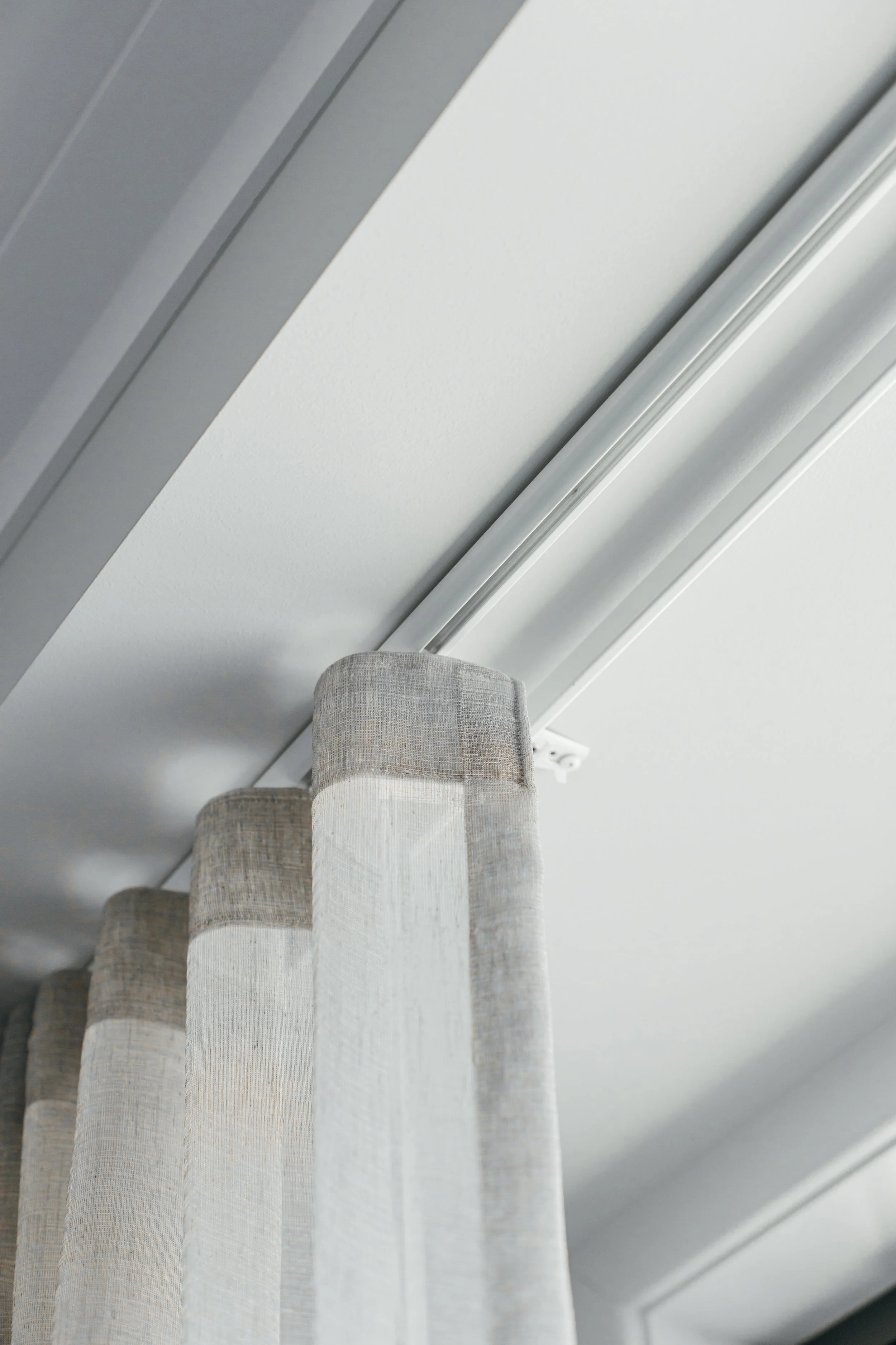 Close-up view of beige and light gray window curtains hanging in front of a white ceiling with molding, viewed from below.