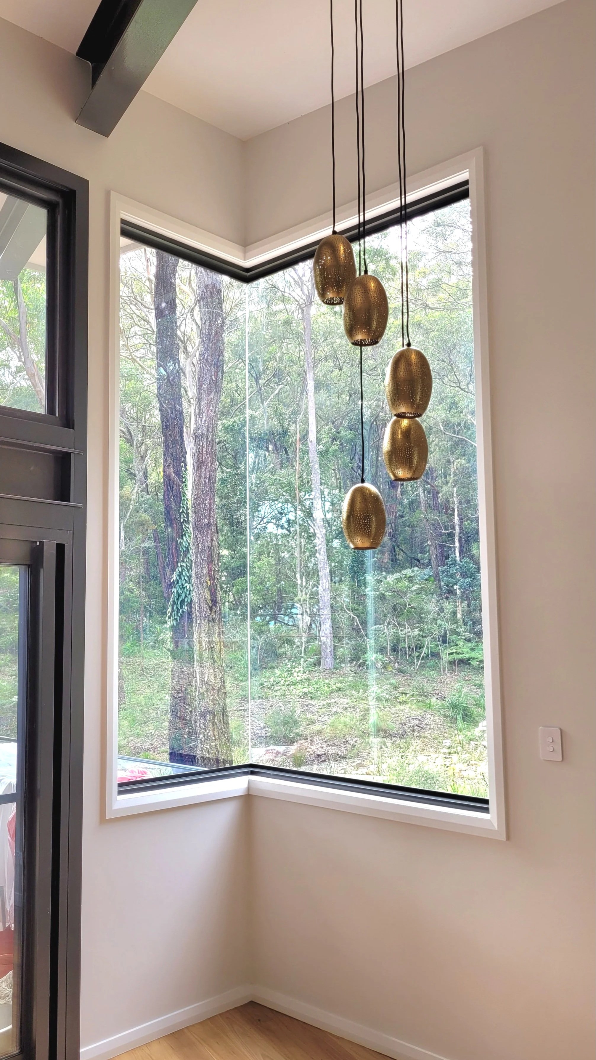 Interior corner of a room with large window showing trees outside, modern hanging pendant lights with gold perforated covers, white walls, and hardwood floor.