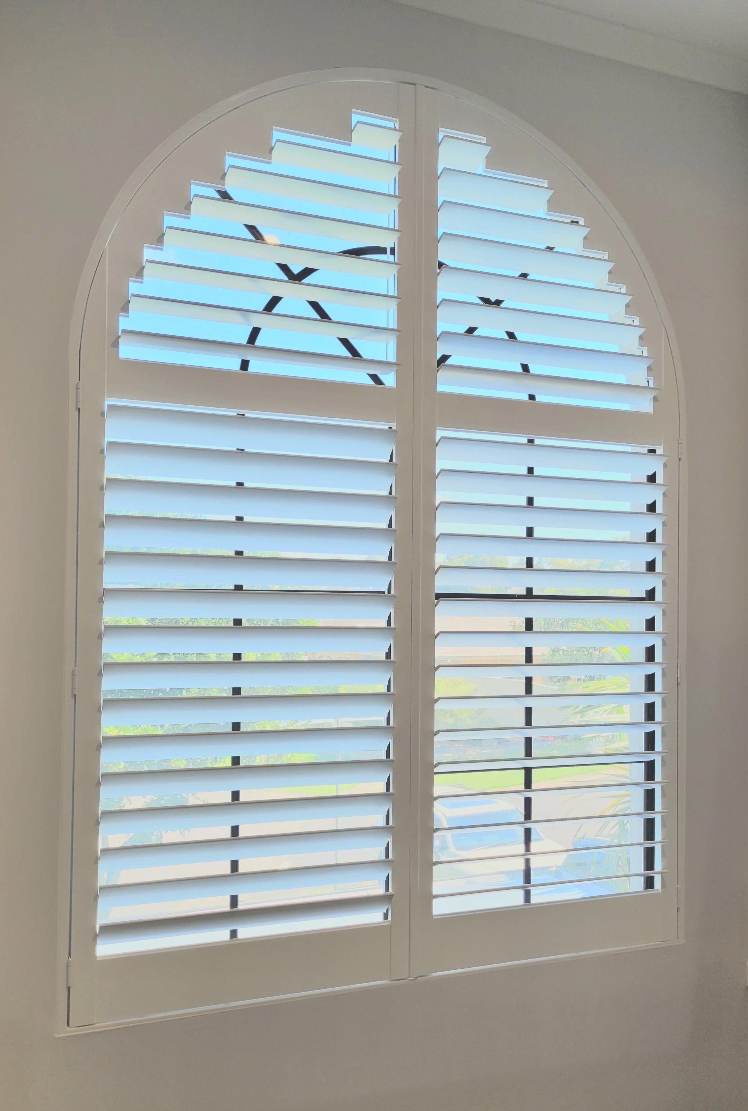 A pair of white plantation shutters with horizontal slats on an arched window, showing a view of a bright outdoor scene with blue sky and greenery.