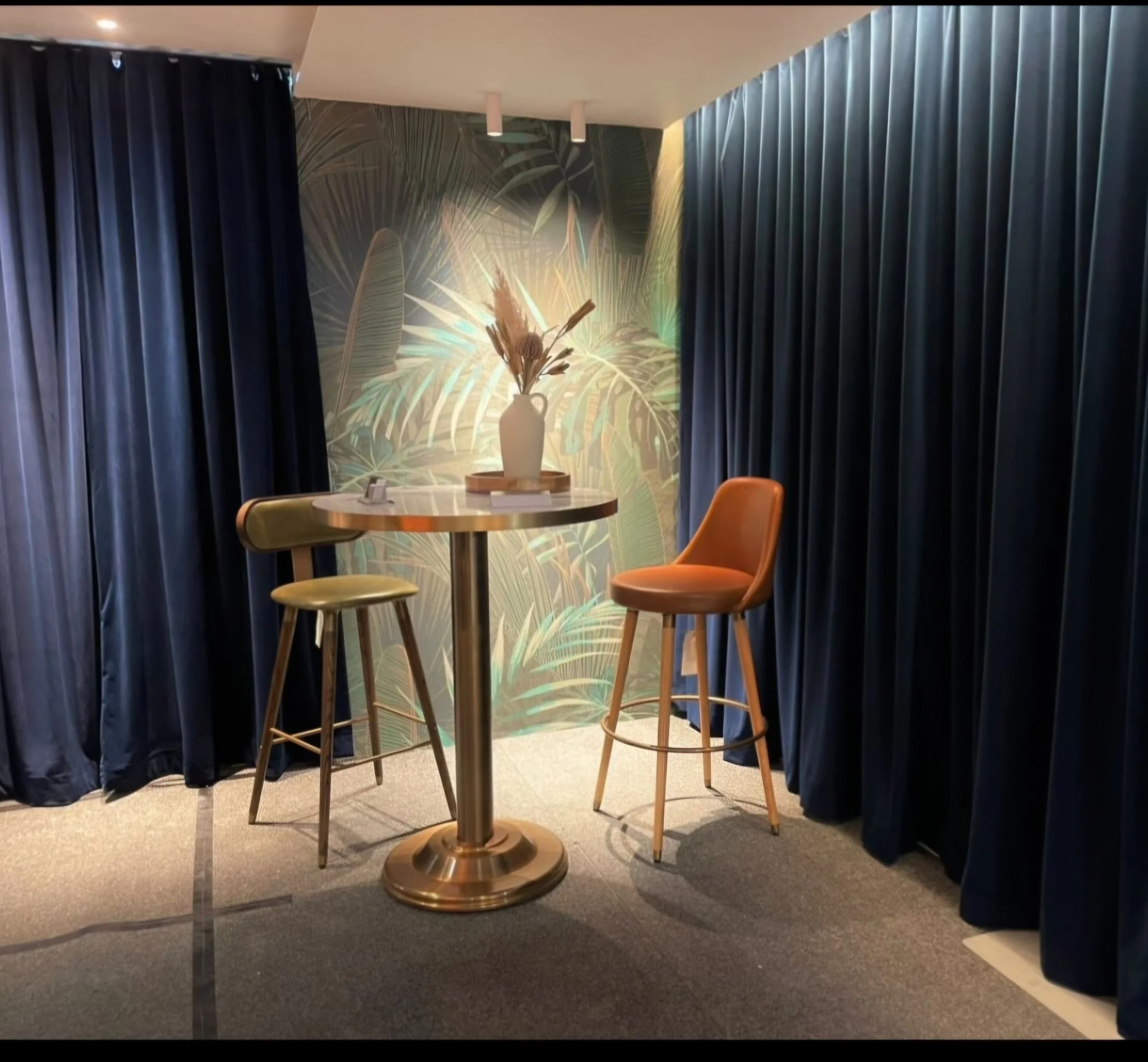 Indoor scene with a high round table, two orange chairs, and a decorative wallpaper featuring large tropical leaves. Dark curtains are on either side, and a vase with dried plants is on the table, lit by ceiling spotlights.